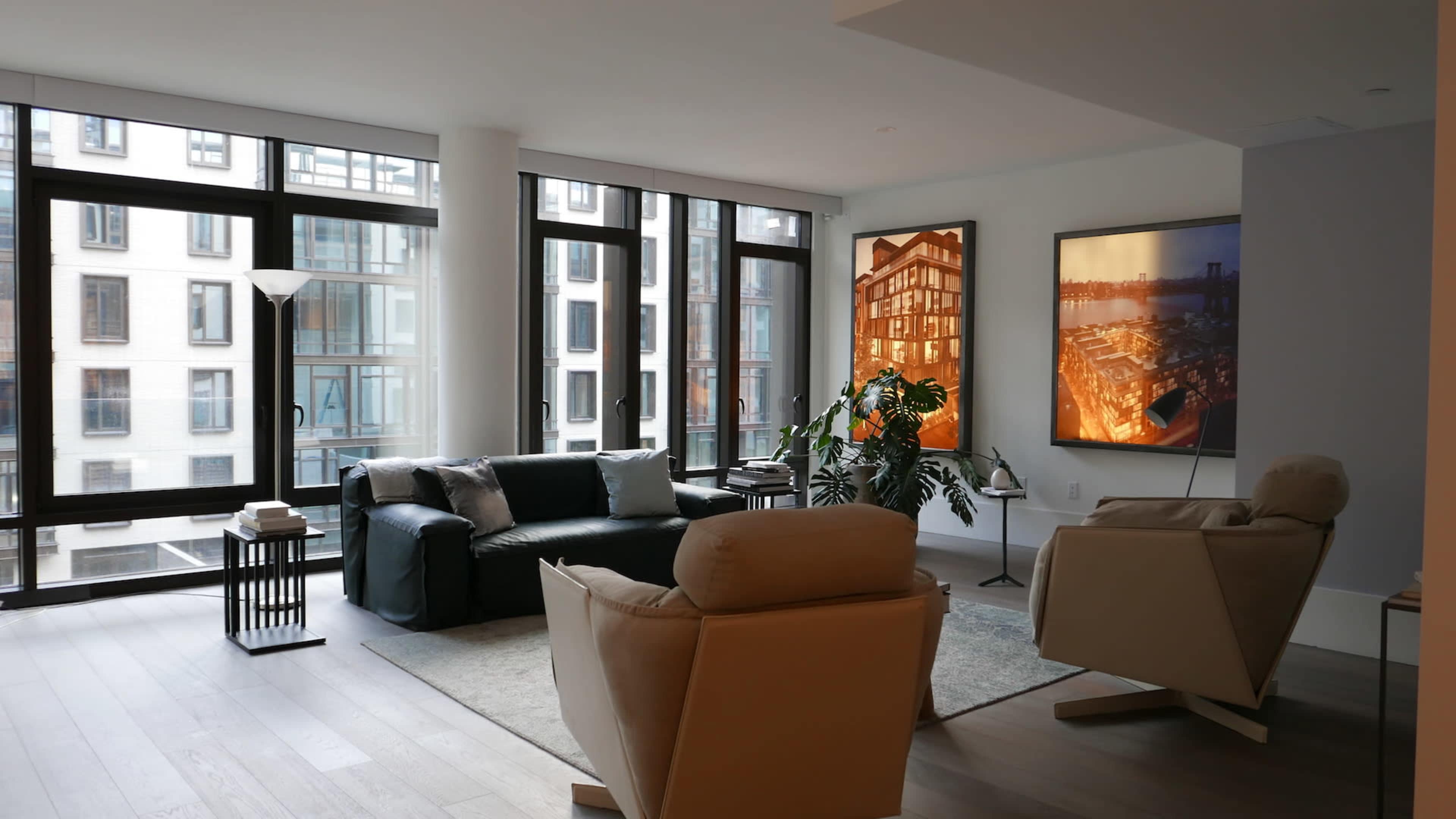 A spacious living room features large windows, modern furniture, and two pieces of artwork on the walls.