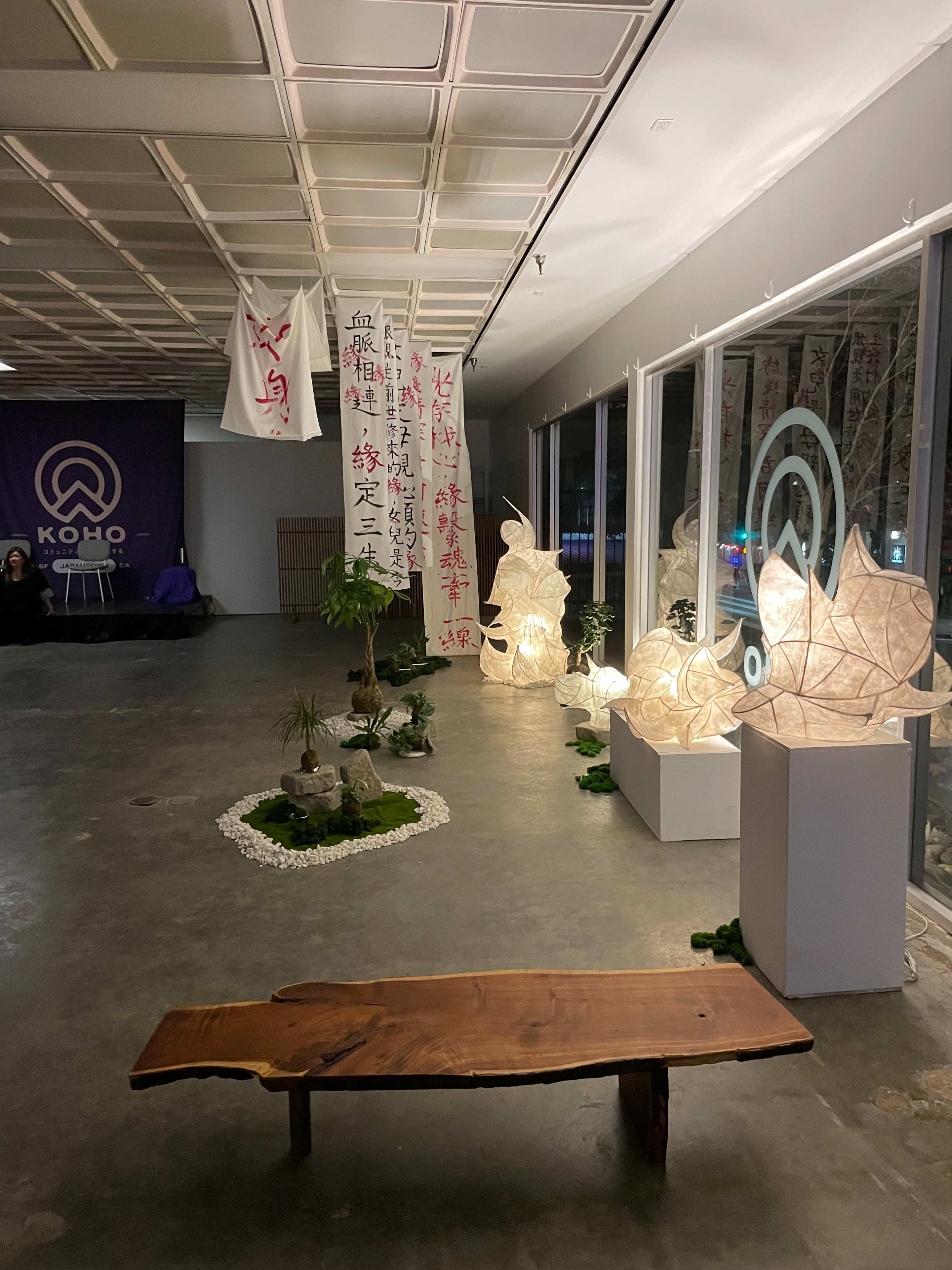 The image shows a contemporary indoor space featuring decorative lanterns, a small plant arrangement, calligraphy scrolls hanging on the walls, and a wooden bench.