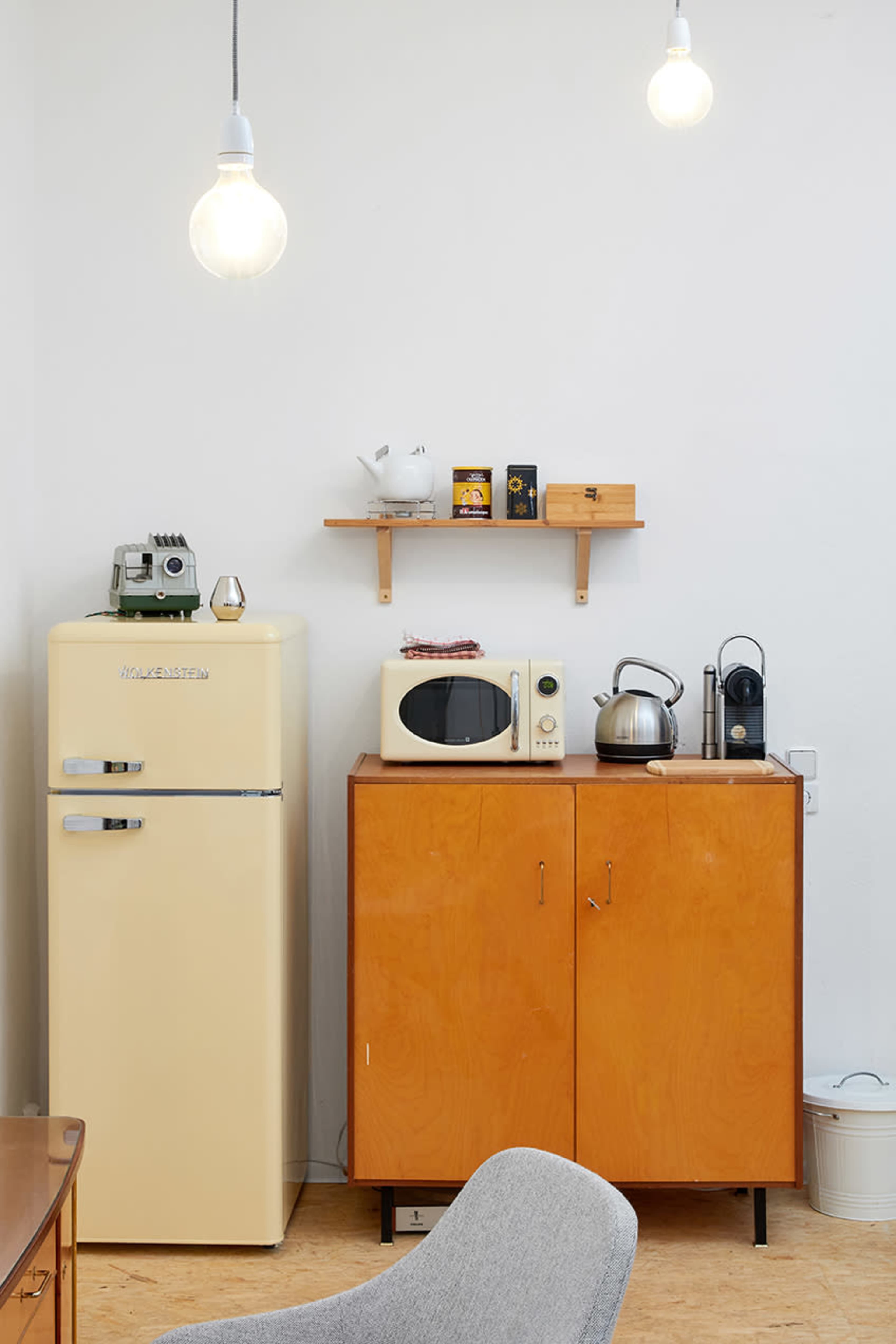 A vintage cream-colored refrigerator and wooden cabinet are positioned against a white wall, along with a microwave, kettle, and various kitchen items on shelves above.