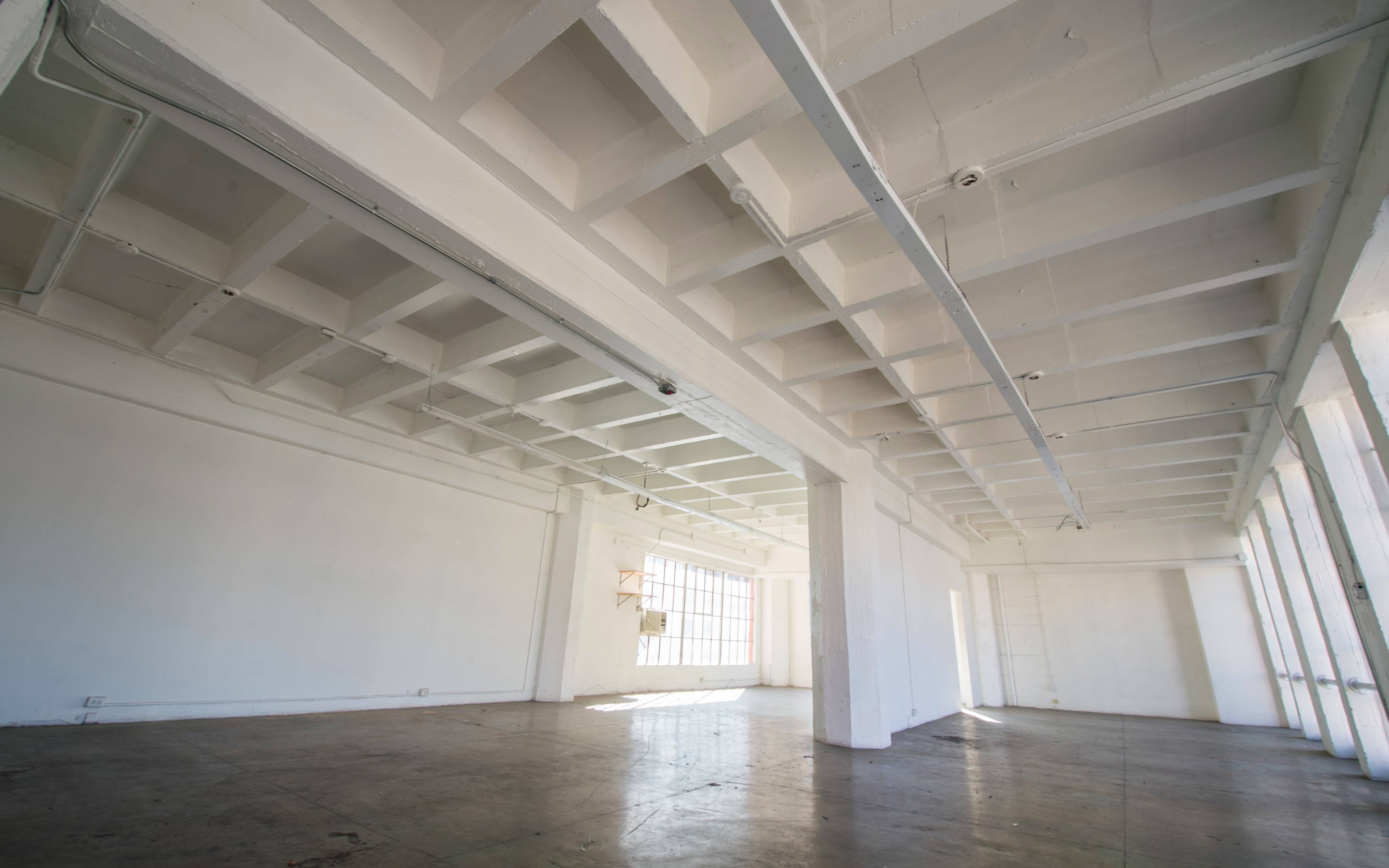 A spacious, empty room with white walls and a coffered ceiling, featuring large windows that allow natural light to enter.