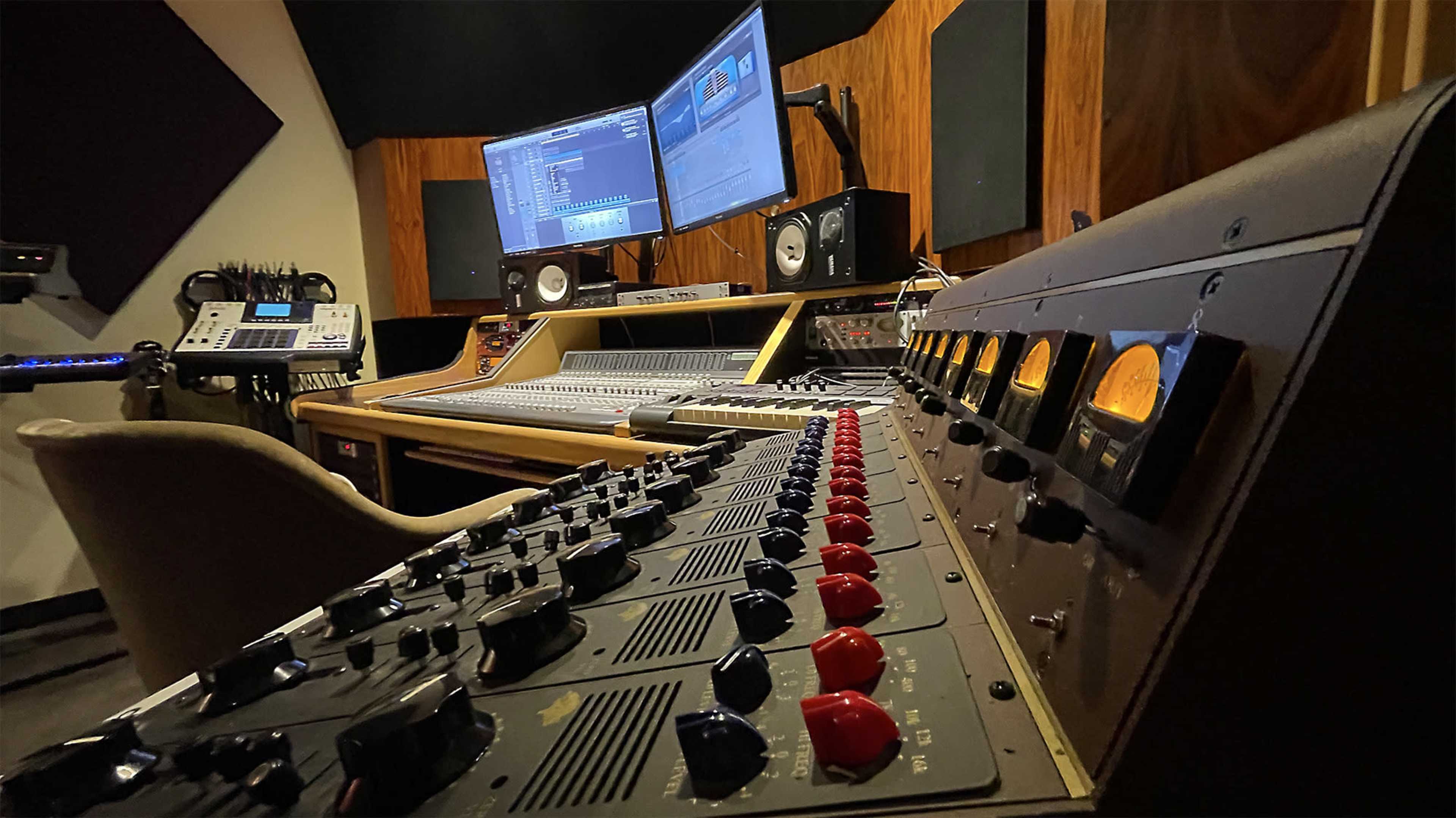 The image shows a recording studio featuring a mixing console with various knobs and faders, two computer monitors displaying audio software, and studio monitors on the shelves.