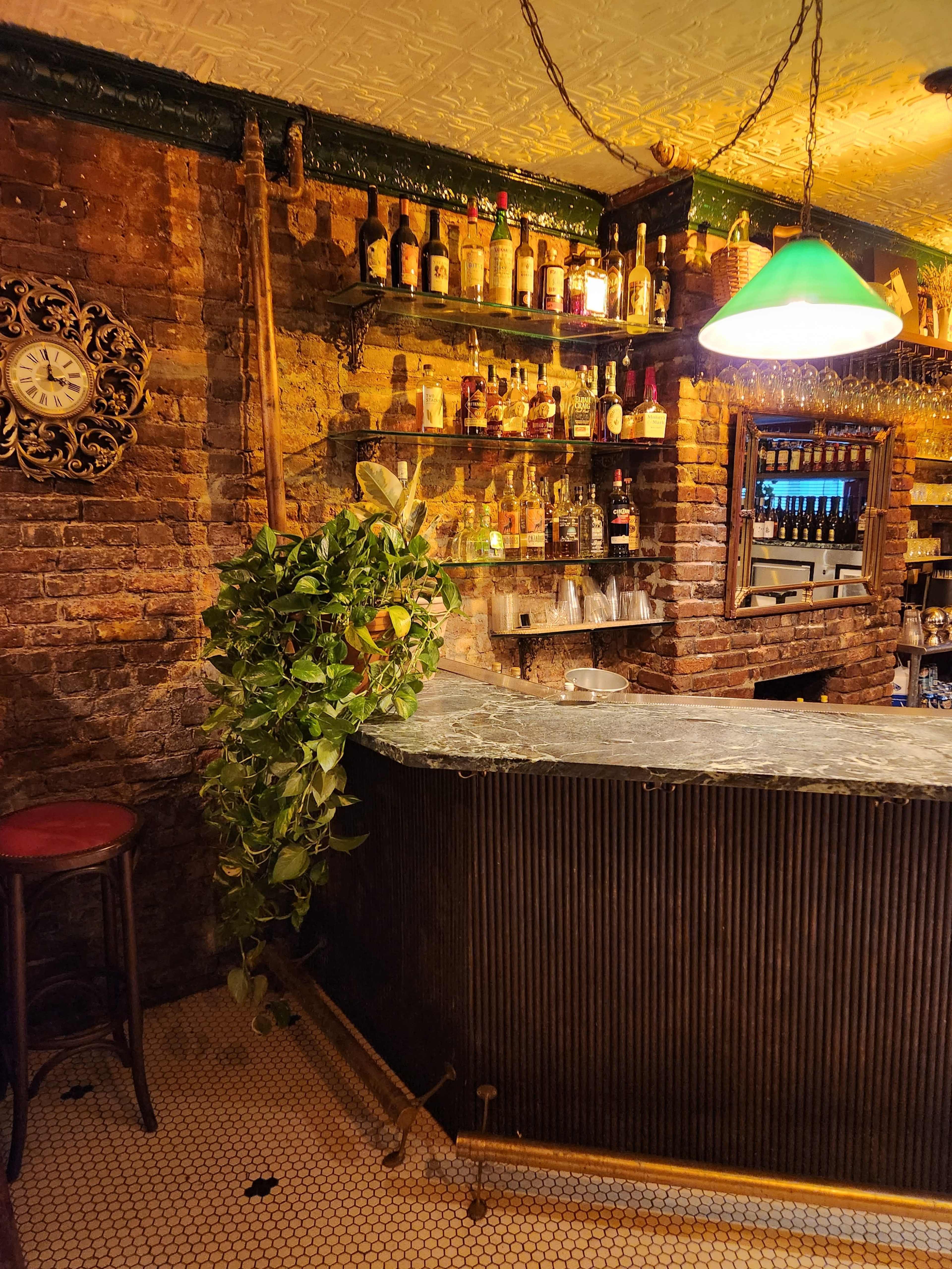 The image shows a rustic bar area with a stone countertop, brick walls, shelves lined with various bottles, and a green lamp overhead.