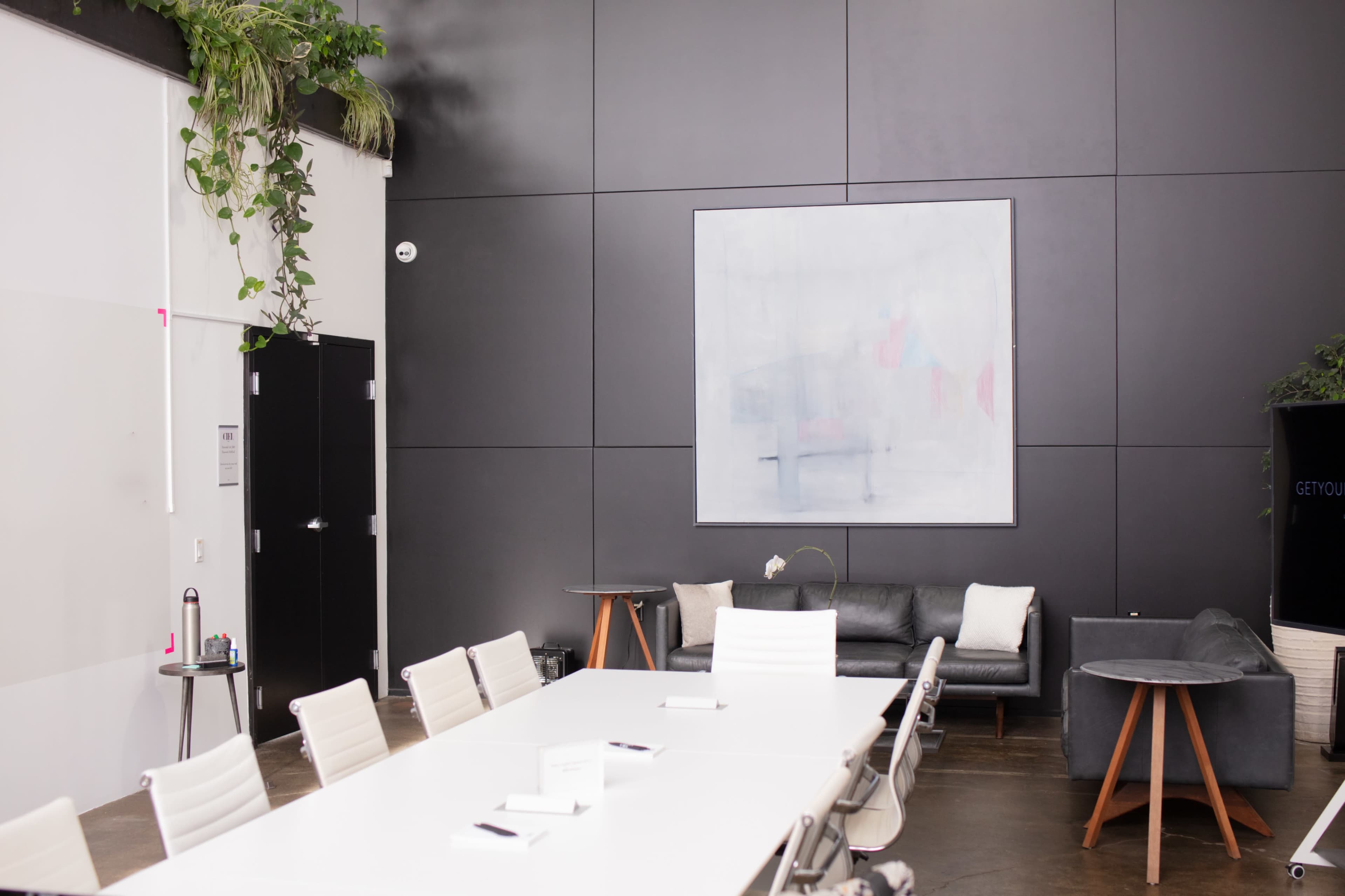 A modern meeting room featuring a large table surrounded by white chairs, a black wall with an abstract painting, and a gray sofa placed next to a green indoor plant.