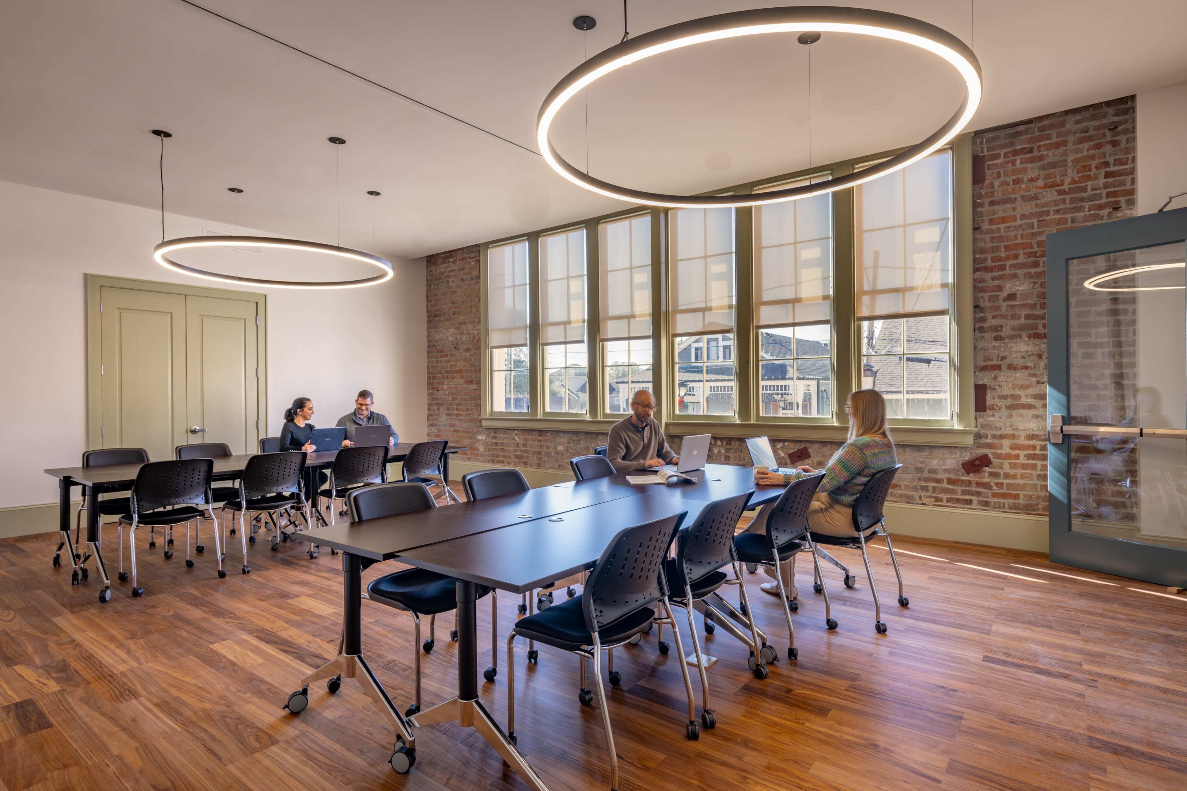 A modern conference room features a large table with several chairs, bright natural light coming through large windows, and a sleek design with two circular light fixtures overhead.