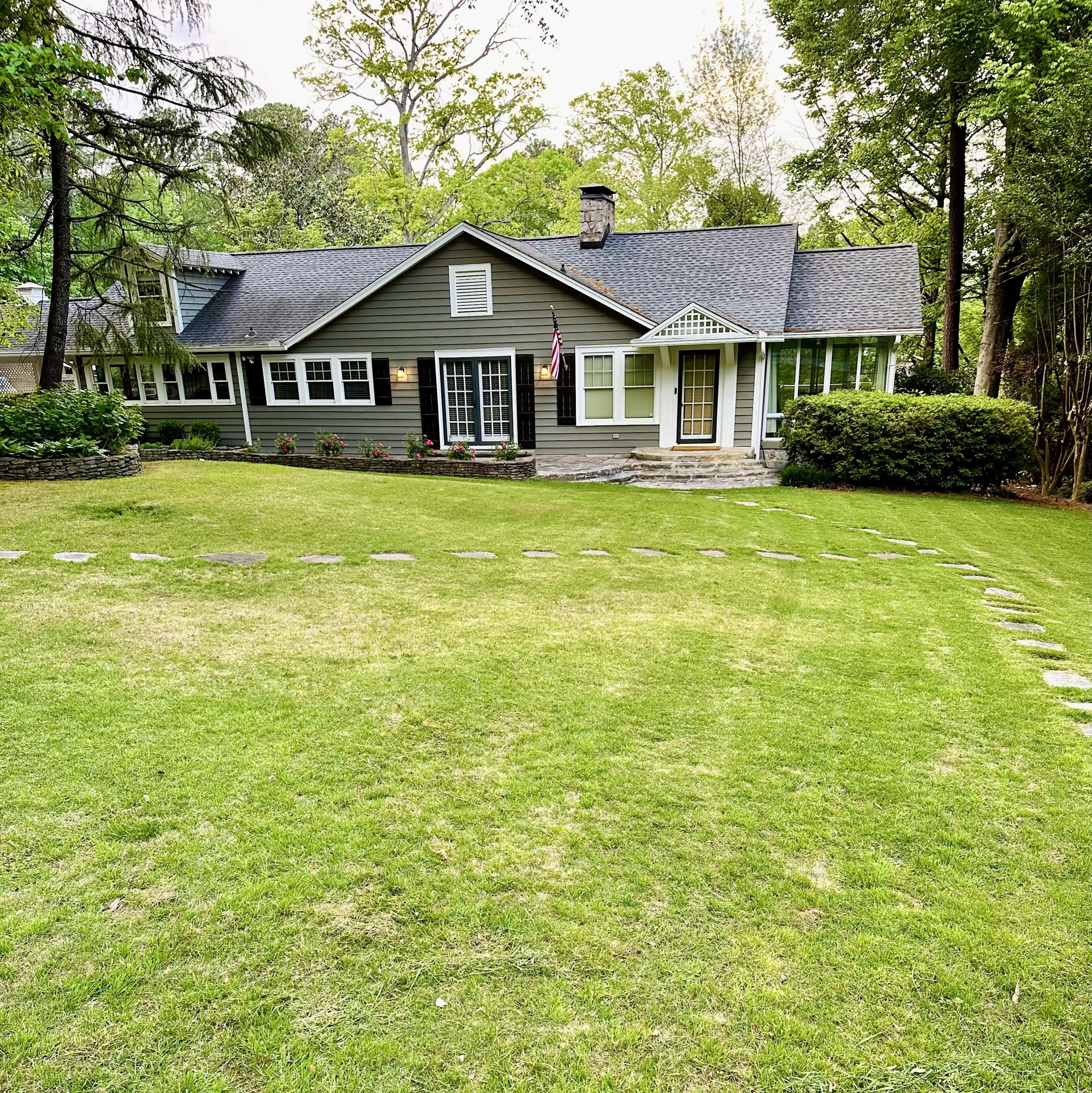 A gray, two-story house with white trim is set amid a lush green lawn, featuring a stone pathway leading to the entrance.