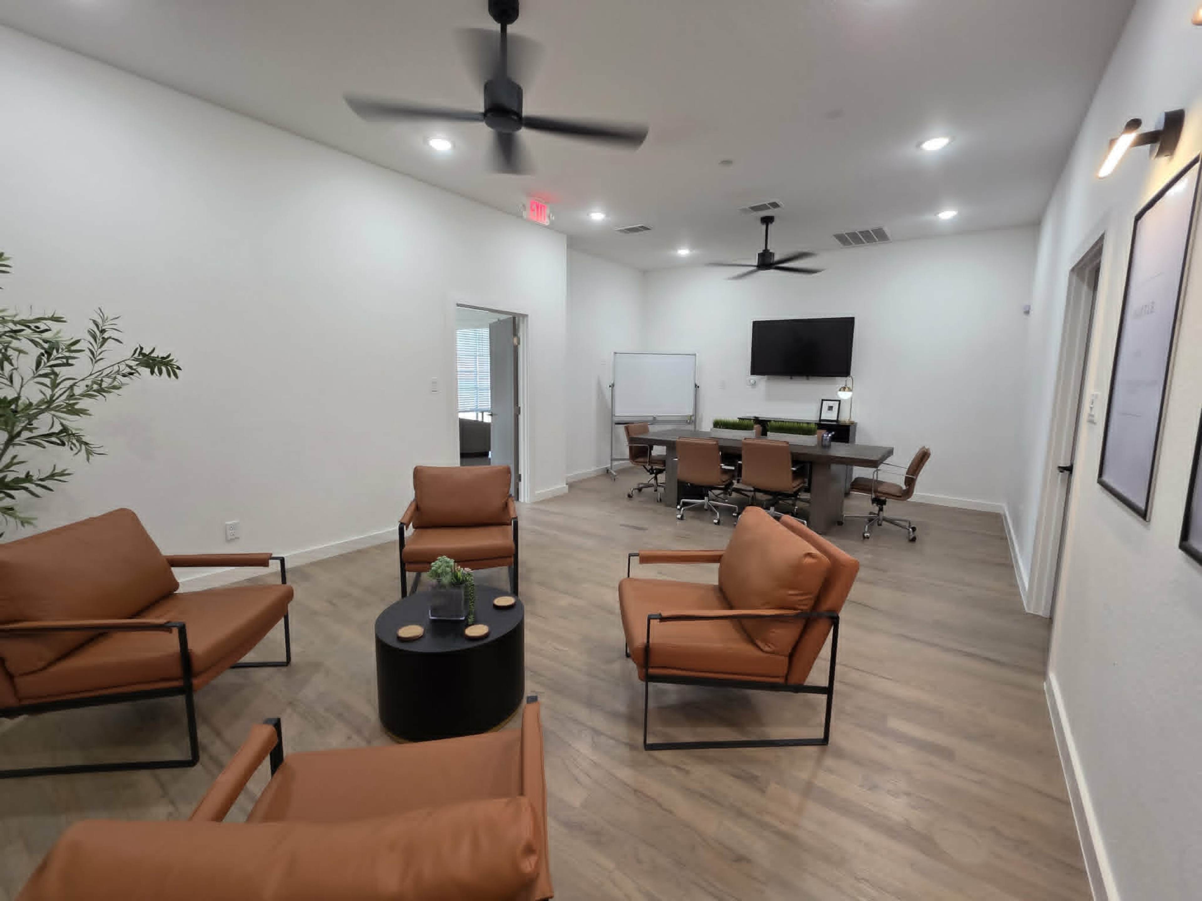 The image shows a modern office space featuring a seating area with four brown chairs around a small black table, and a conference room equipped with a large table and a TV screen.