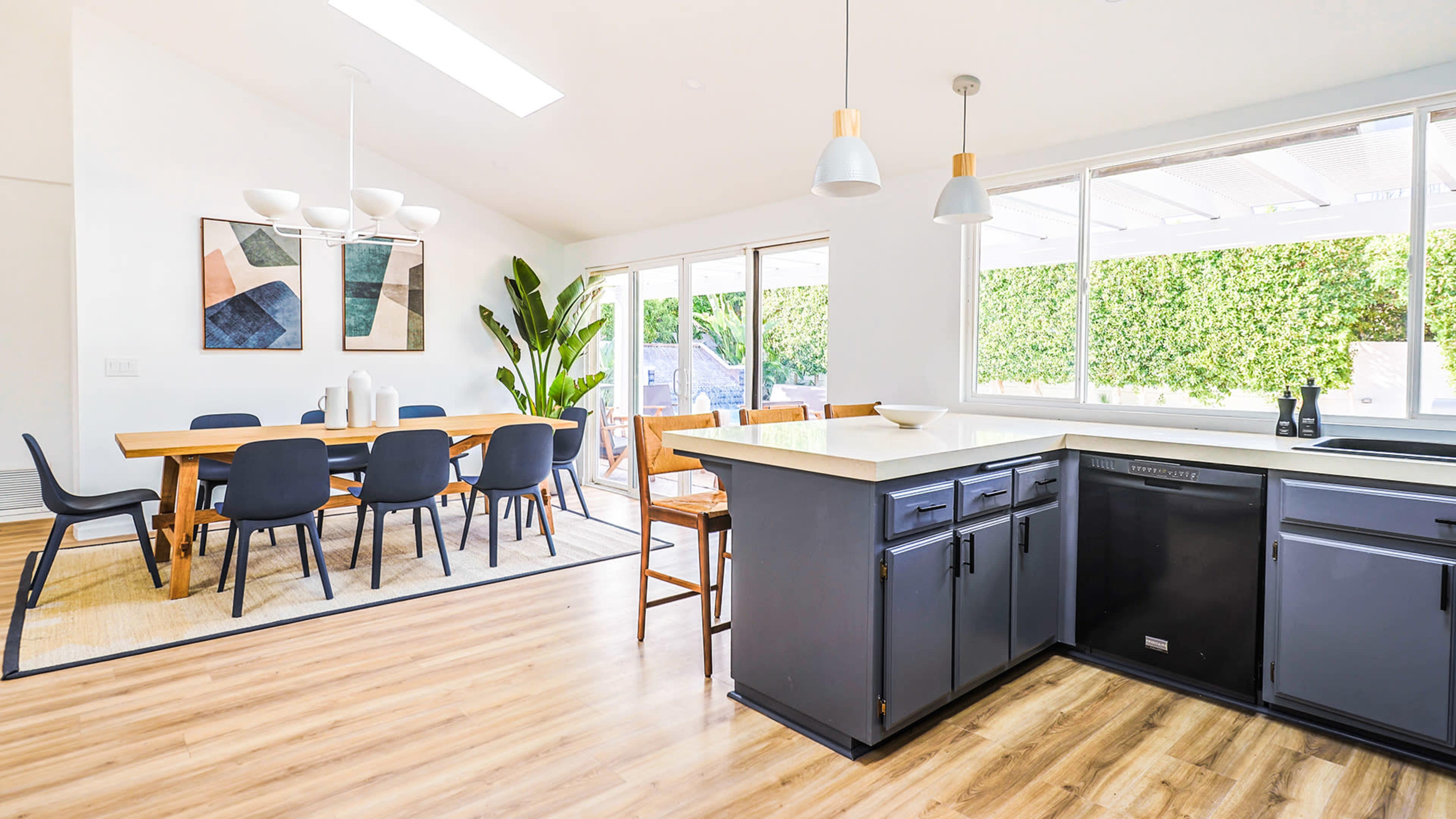 The image shows a bright, modern kitchen connected to a dining area with a large table and sliding glass doors leading to an outdoor space.