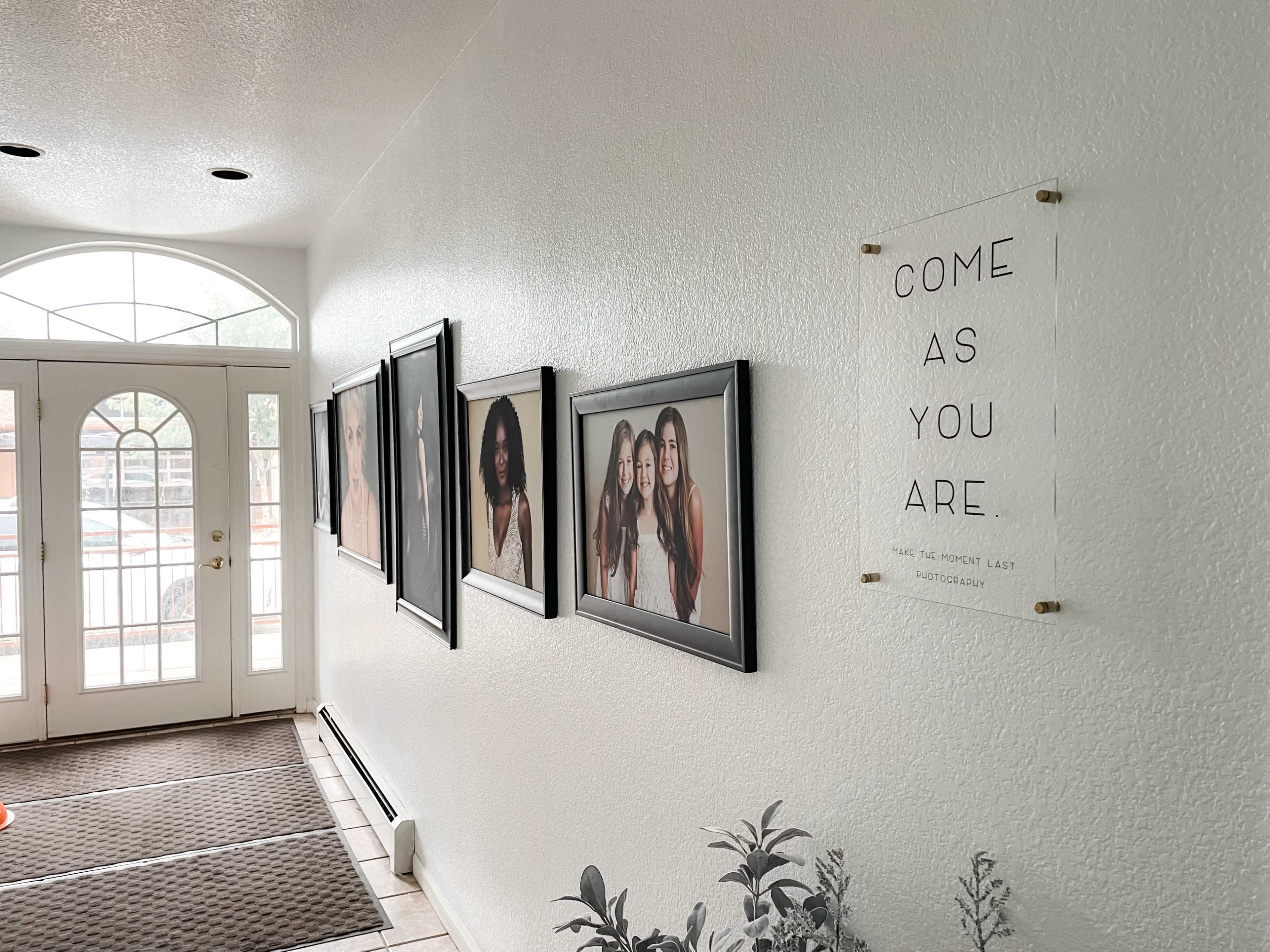 A hallway featuring framed portraits on the wall and a decorative sign that reads "COME AS YOU ARE," with a front door visible in the background.