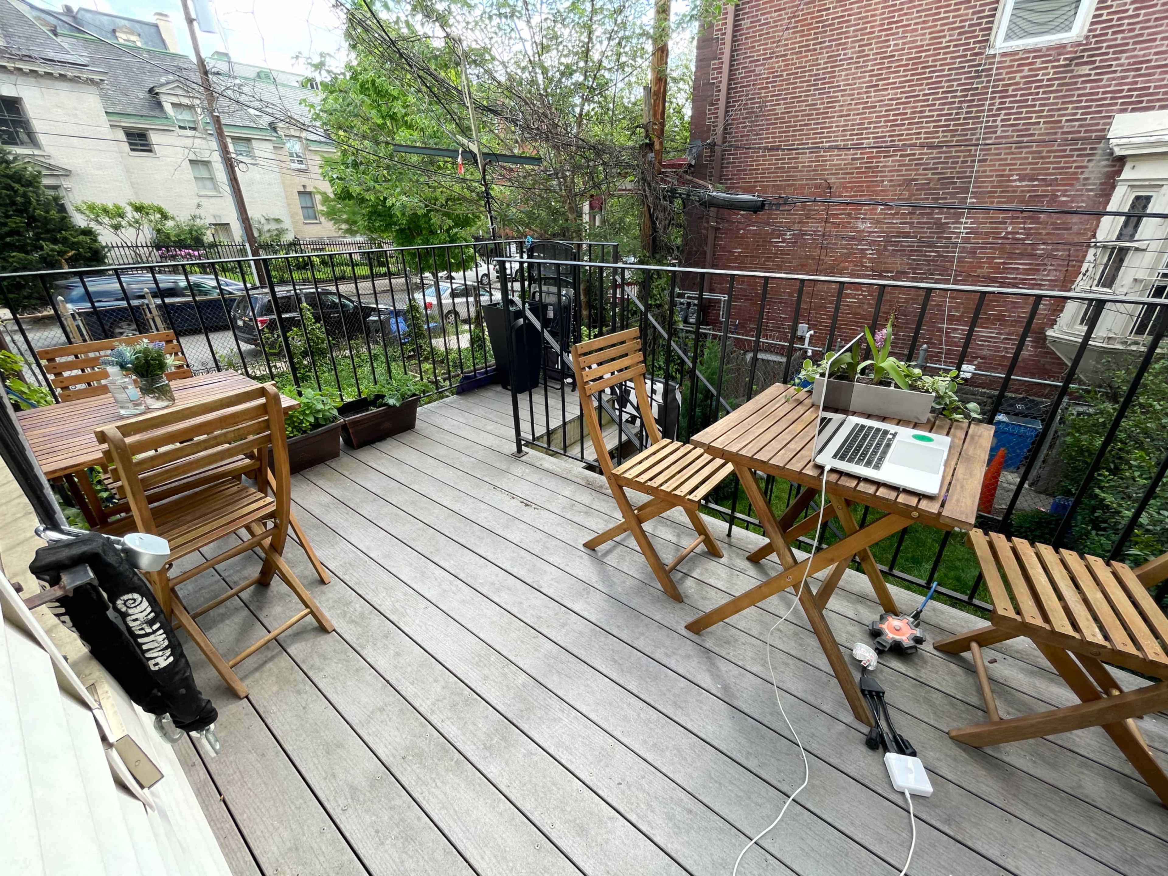 A small wooden deck with two folding tables and chairs, a laptop on one table, and potted plants along the edge.