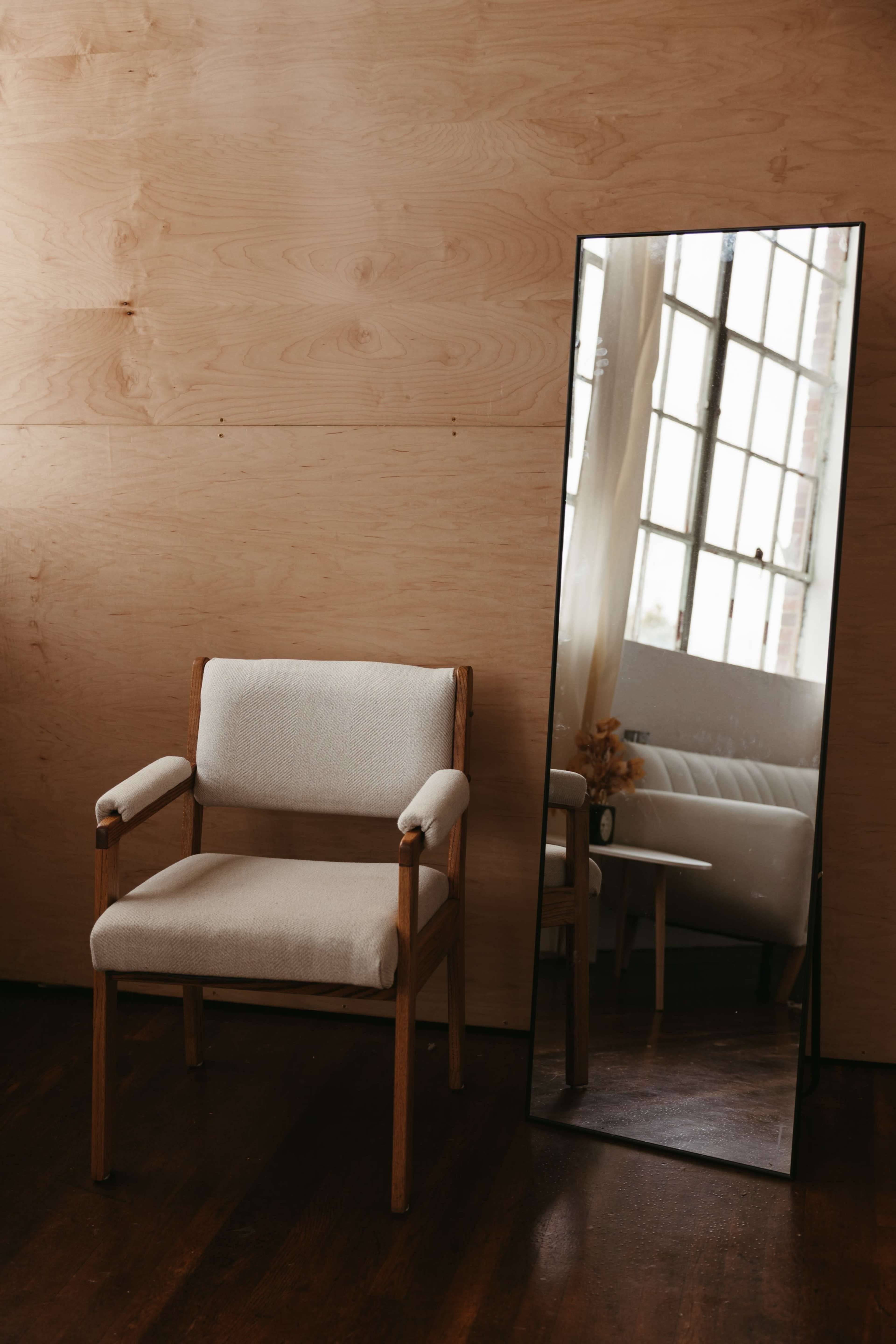 A simple wooden chair is positioned next to a tall mirror reflecting a window and a sofa in a room with wooden walls.