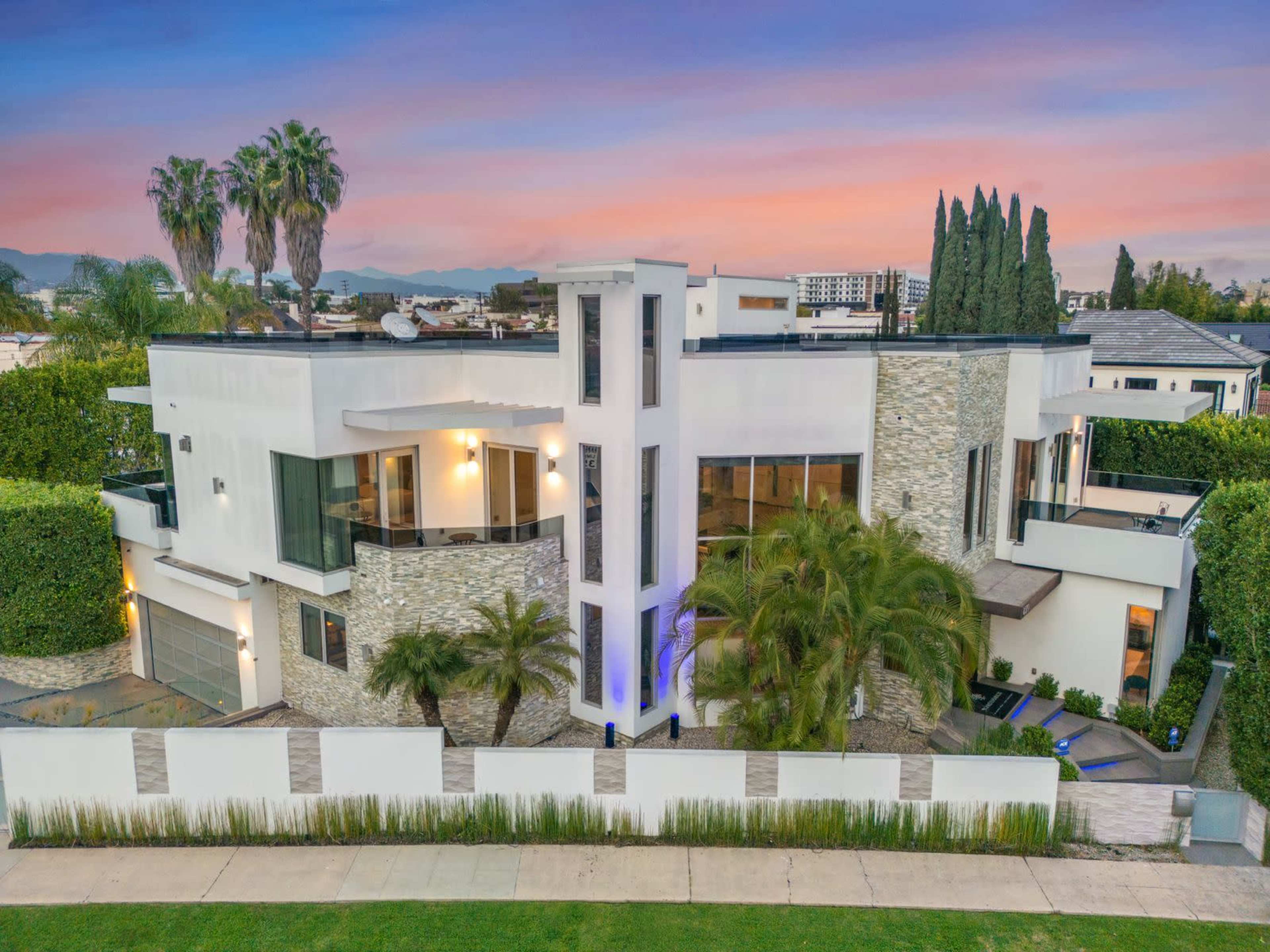 A modern two-story house features a mix of stone and white siding, large windows, and an array of palm trees in the surrounding landscape.