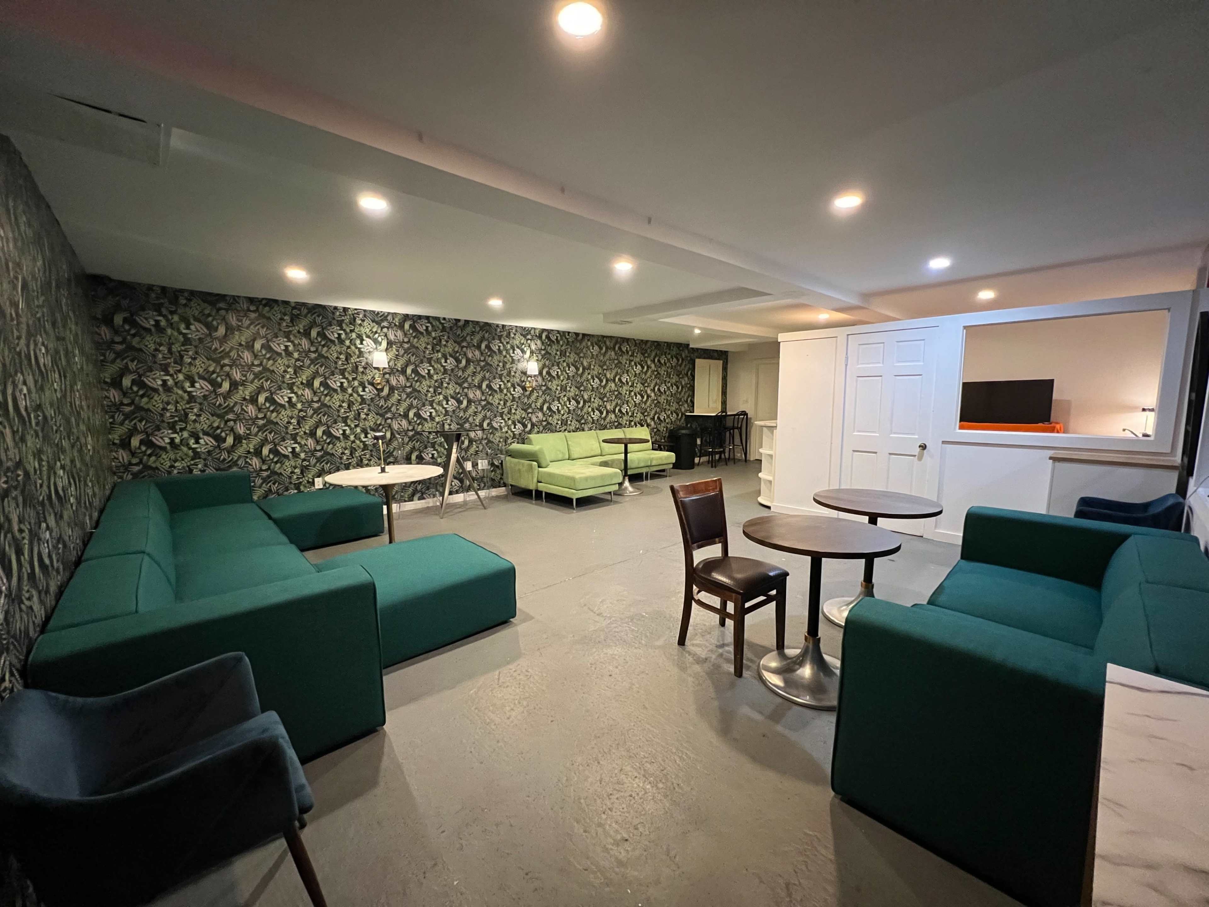 A spacious lounge area with green couches, round and square tables, and a wall featuring a patterned wallpaper.