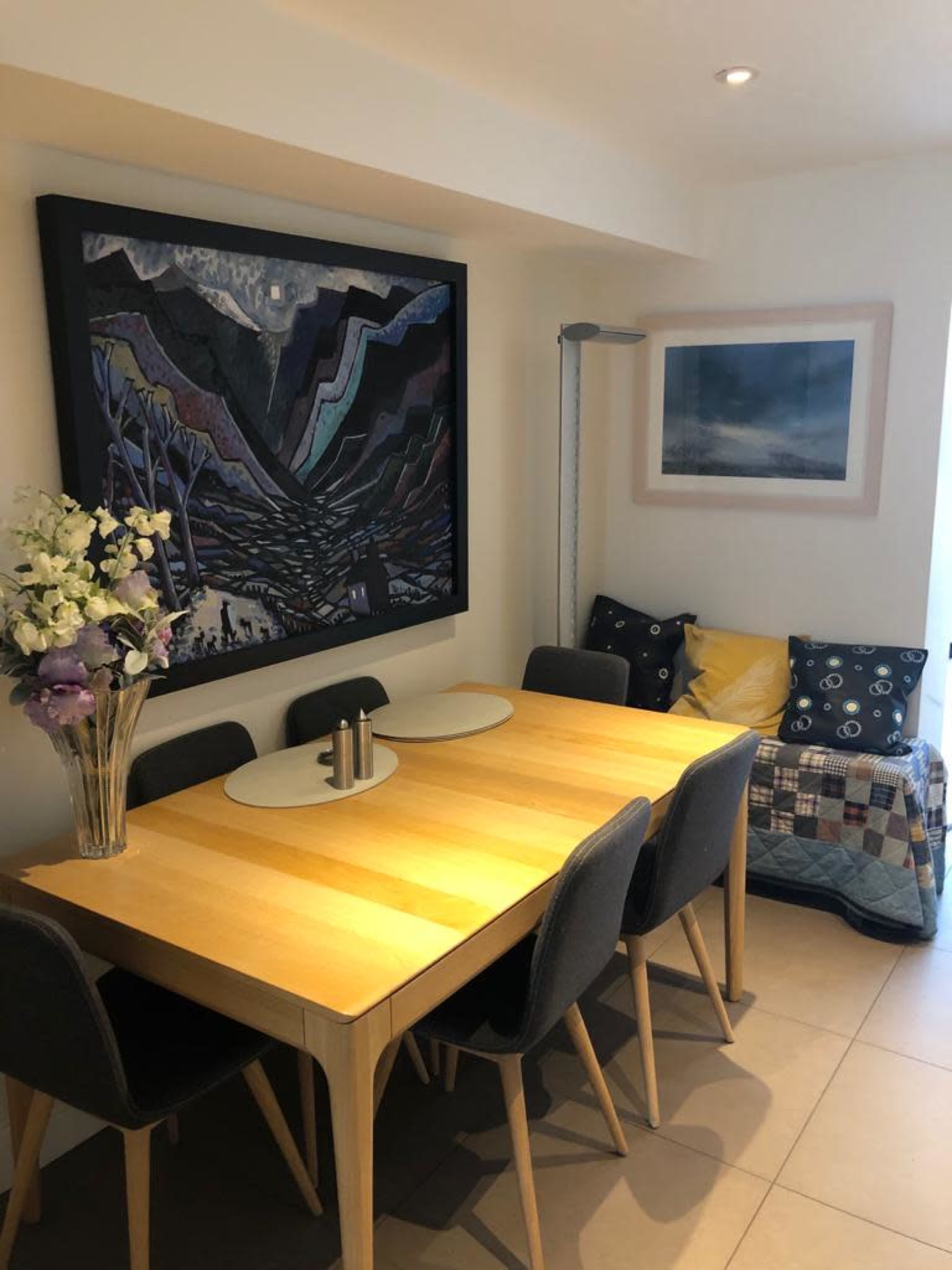 A dining area with a wooden table surrounded by chairs, a large framed artwork on the wall, and a cozy seating nook with cushions.