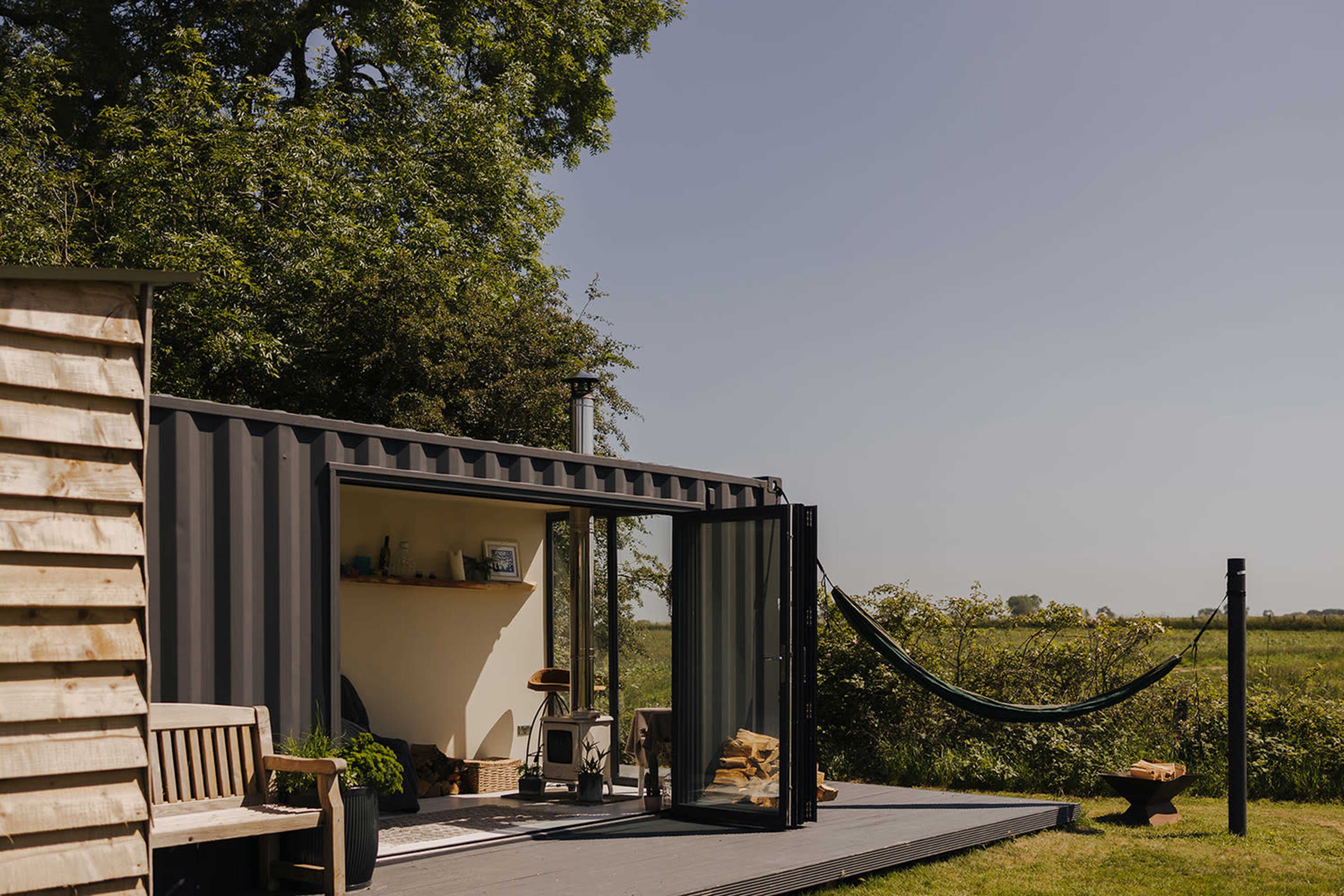 A modern, shipping container-style cabin sits on a deck with a hammock, surrounded by greenery and open fields.