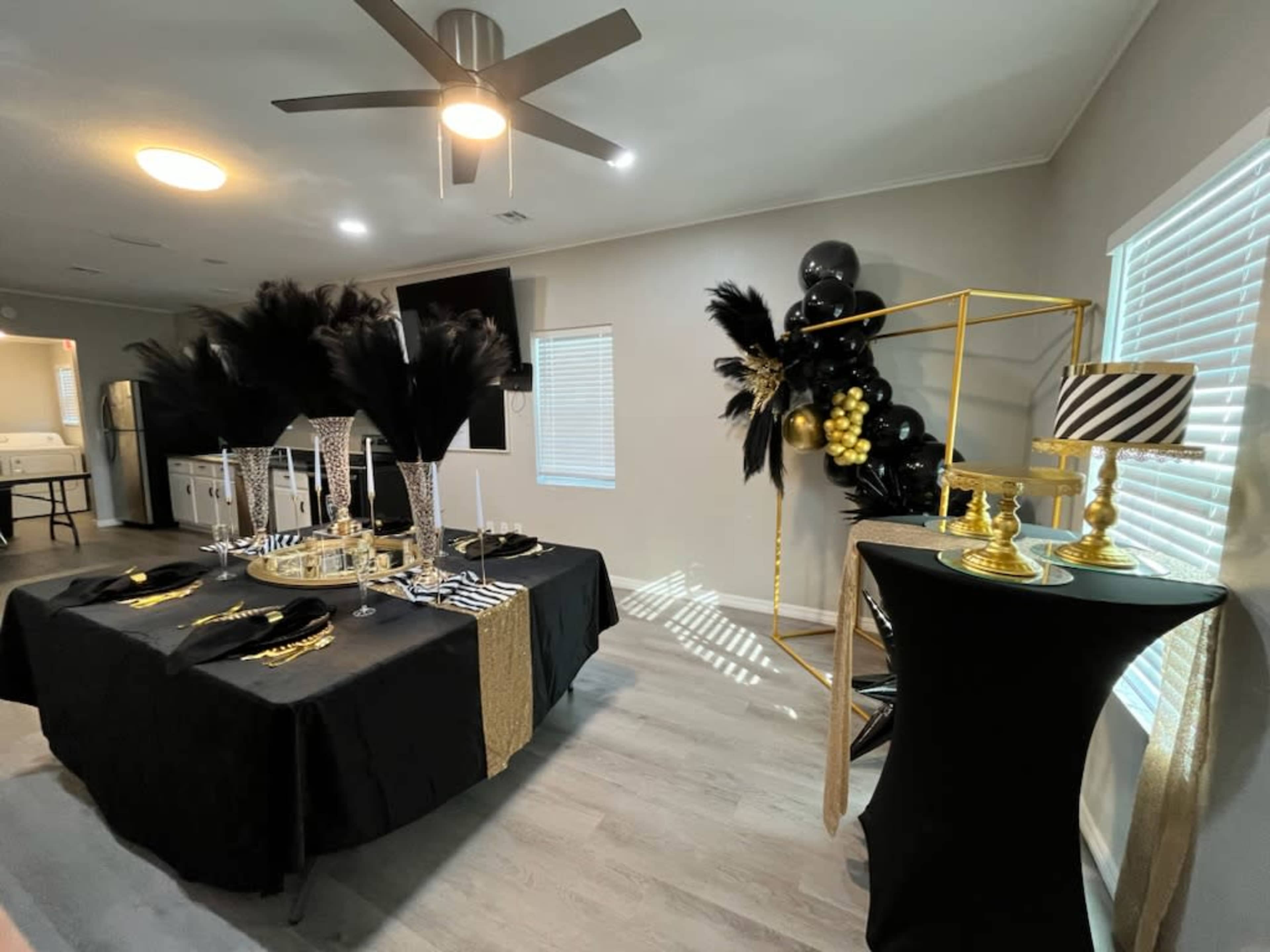 The image shows a decorated interior space with a dining table covered in black and gold accents, featuring elegant tableware and decorative elements, alongside a black and gold balloon arch and a stylish lamp on a separate table.