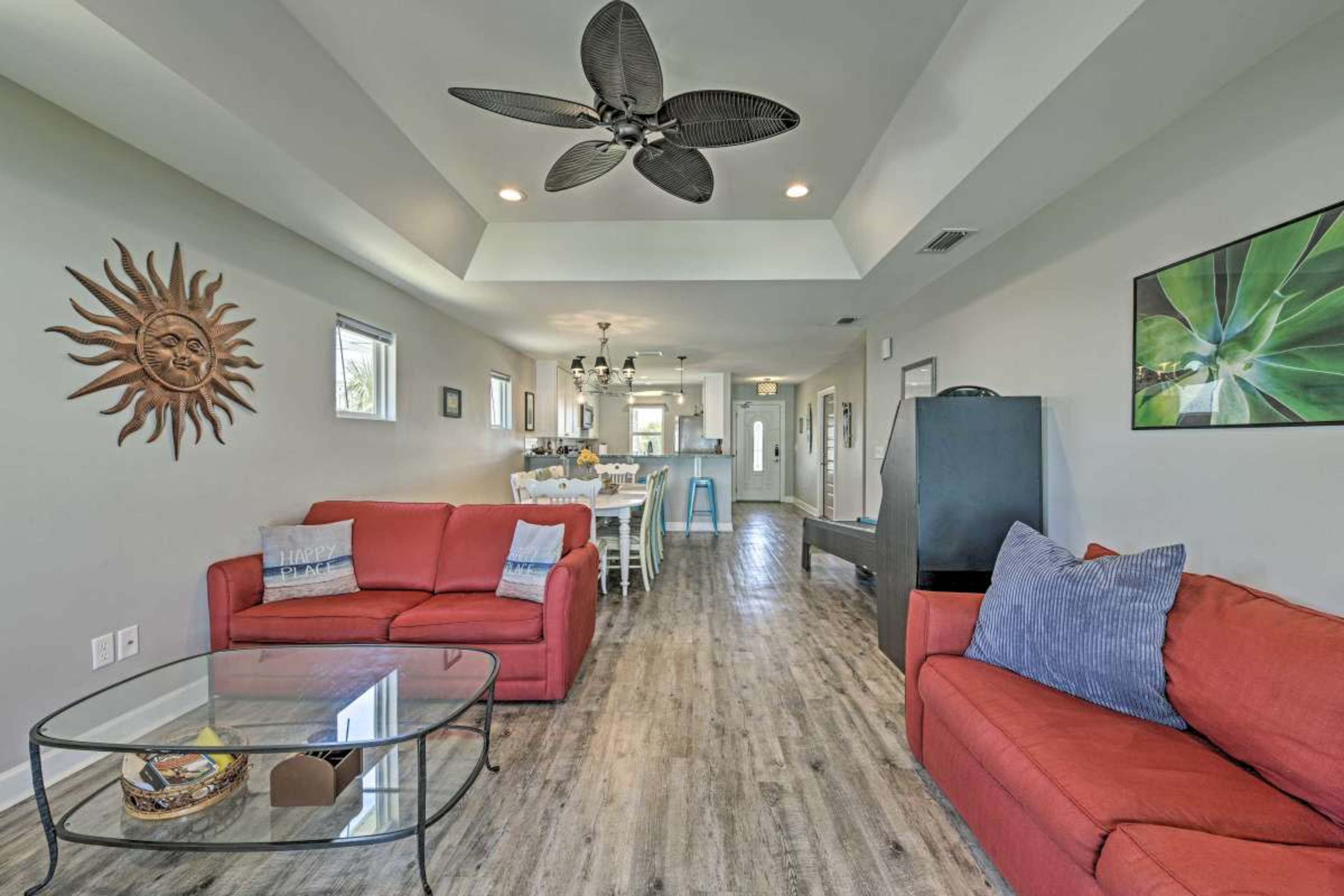 A living area with two red sofas, a glass coffee table, and a ceiling fan, leading into a dining space and a kitchen at the back.