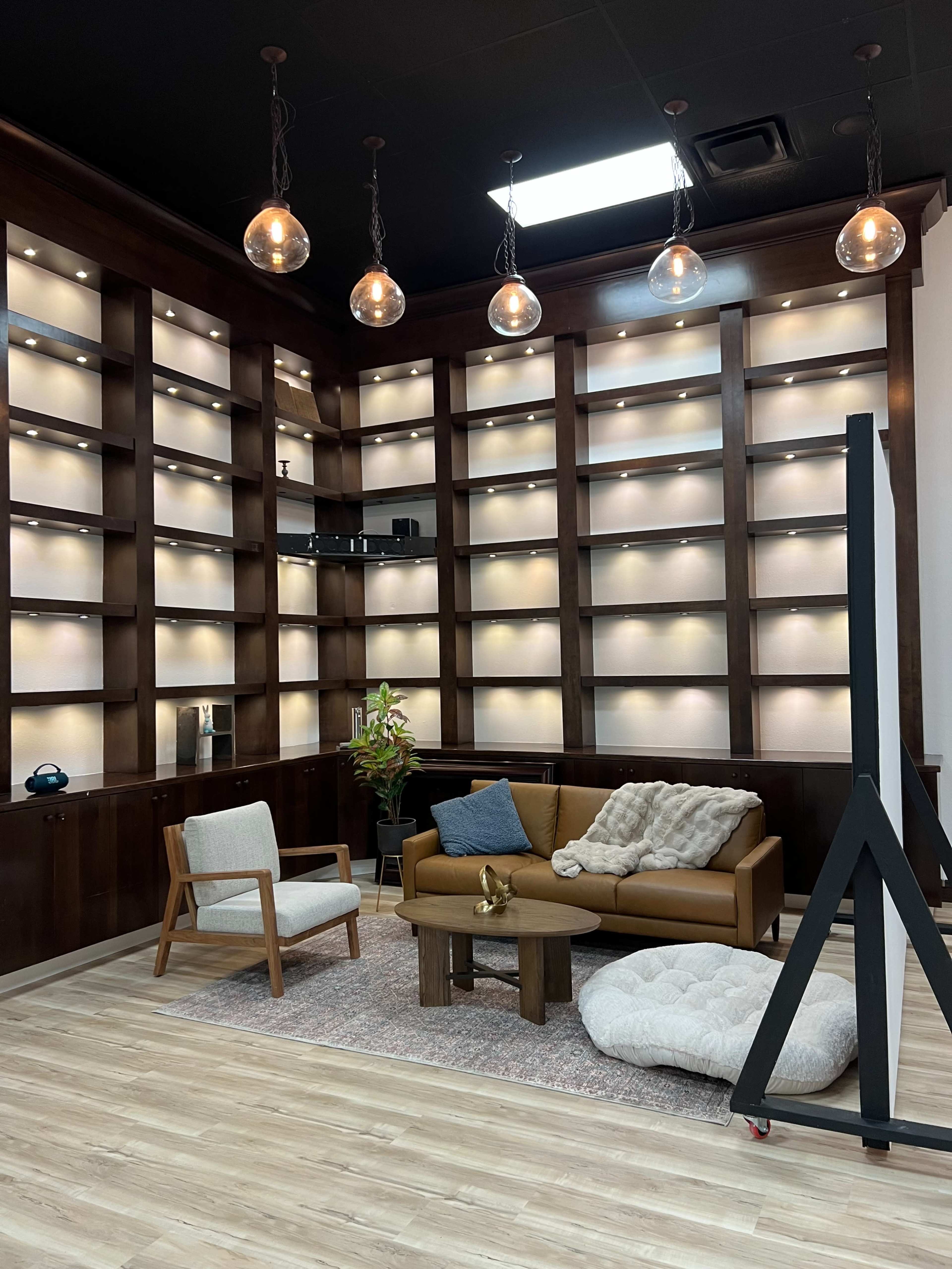 The image shows a modern interior space with wooden shelves illuminated by lights, a brown sofa, an armchair, and a soft floor cushion.