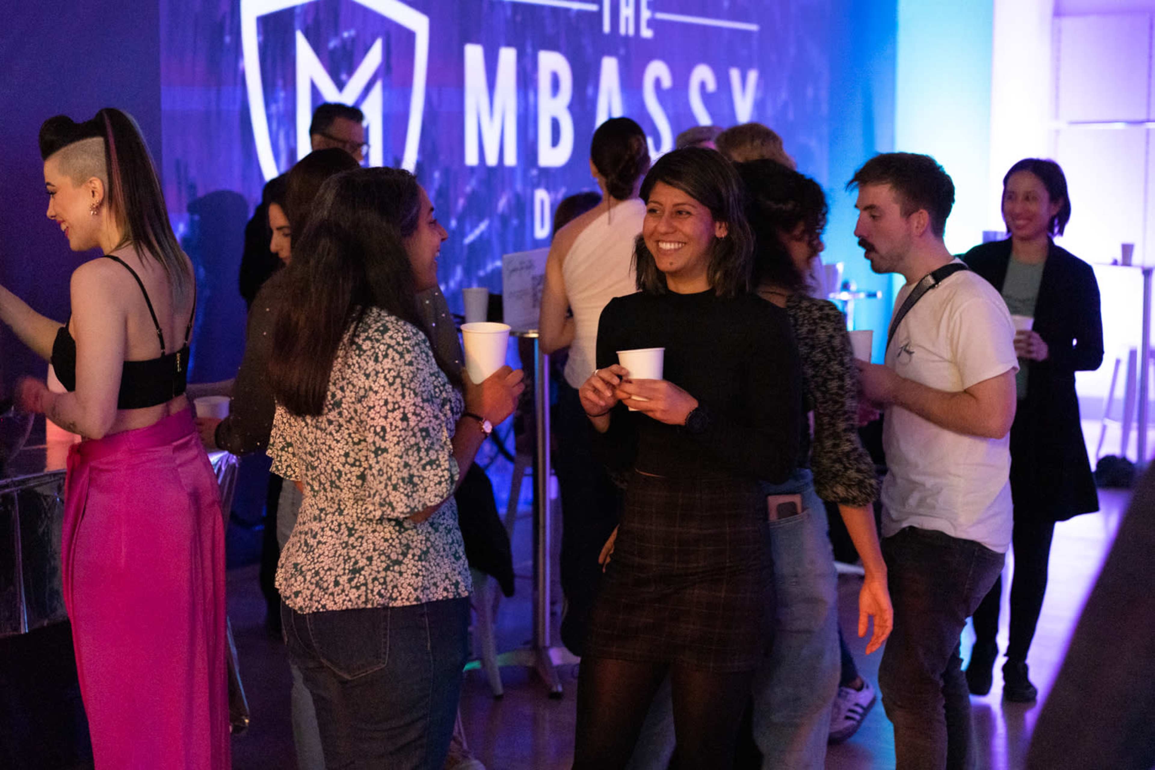 A group of people socialize at an event held at The M-BASSY, with some holding cups and others engaged in conversation.