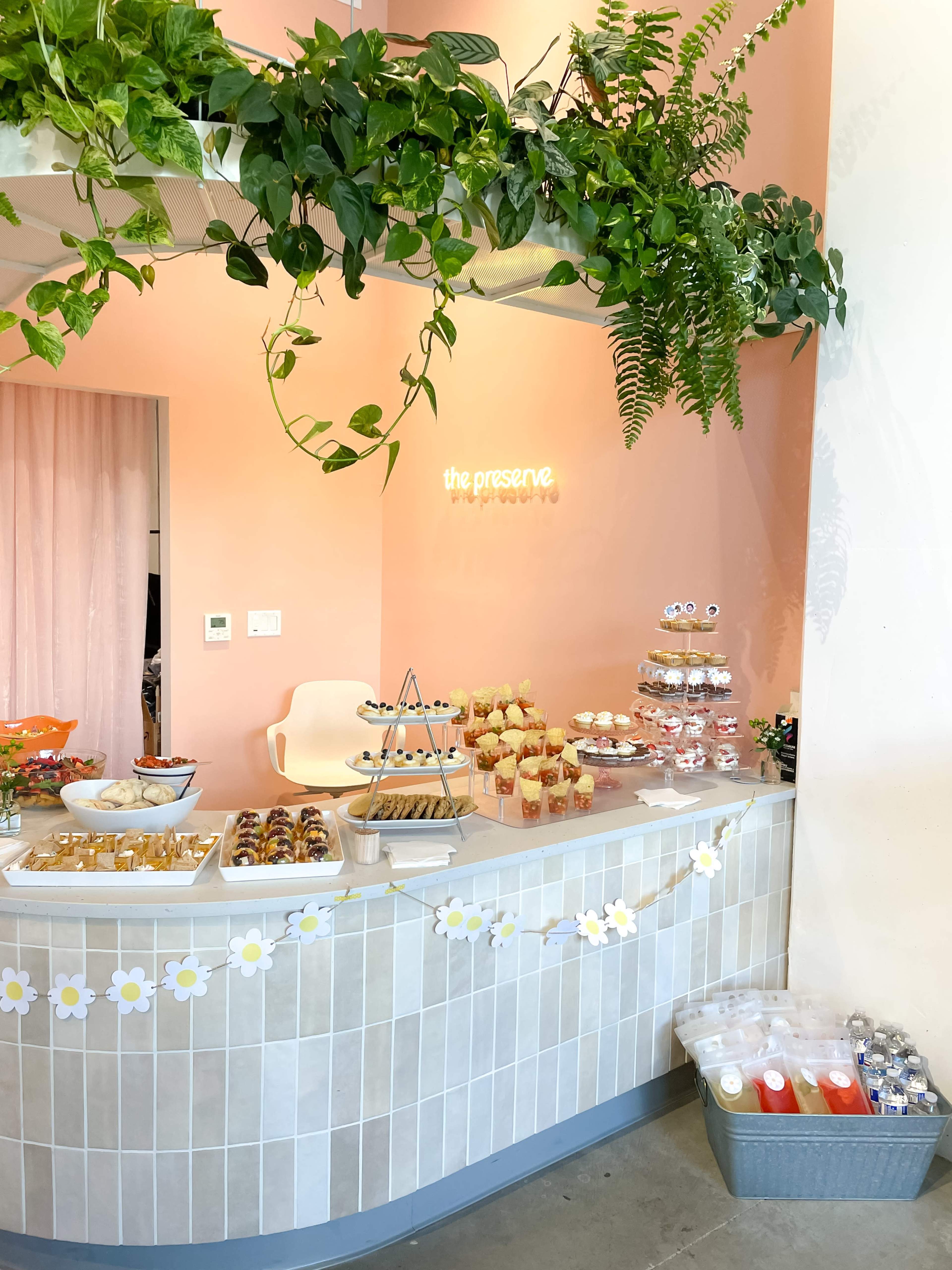 A brightly lit café displays an array of desserts and snacks on a curved countertop, surrounded by green plants and a pastel pink wall.