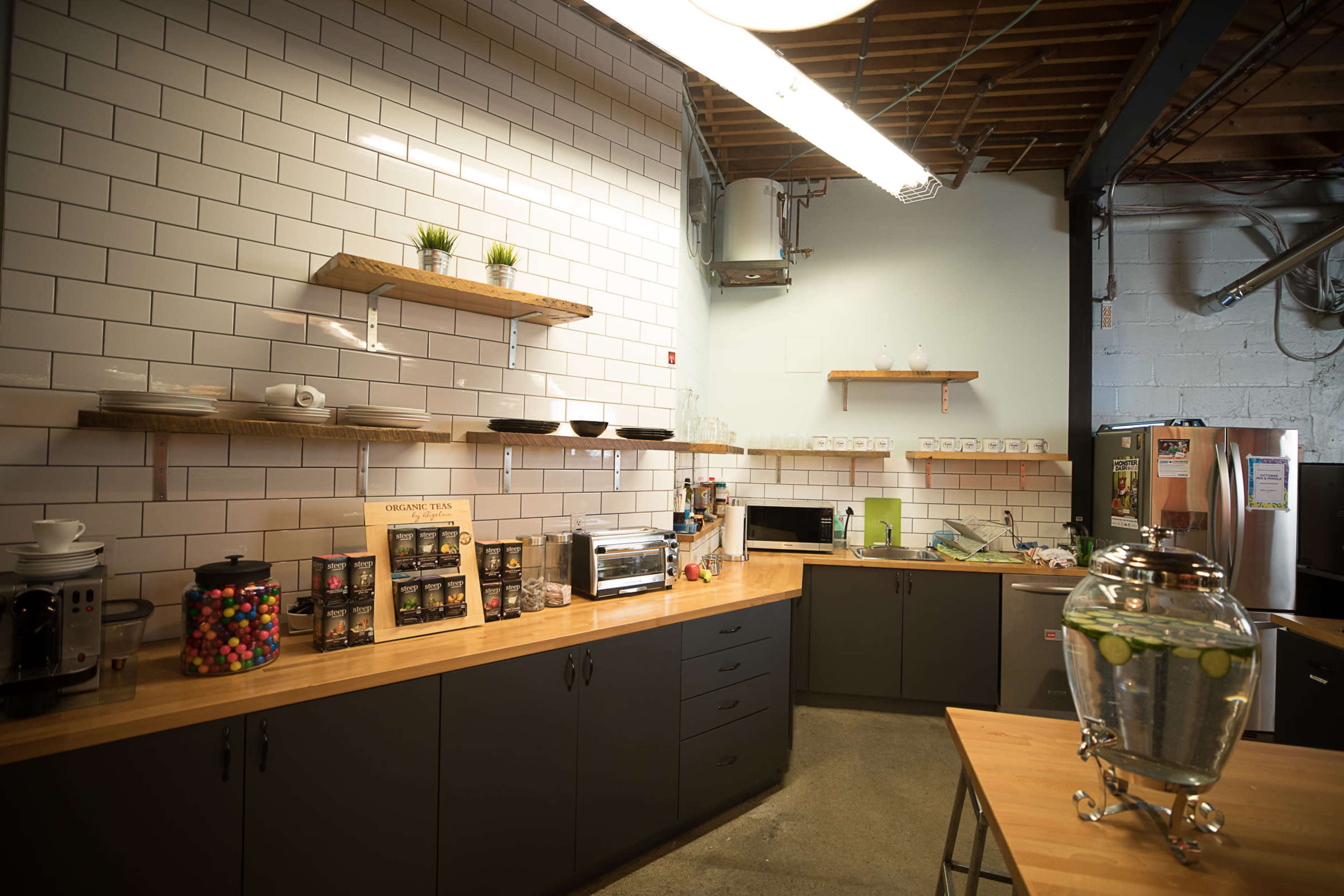 The image shows a modern kitchen with wooden countertops, open shelving, and a variety of appliances, including a coffee maker and a large glass water dispenser.