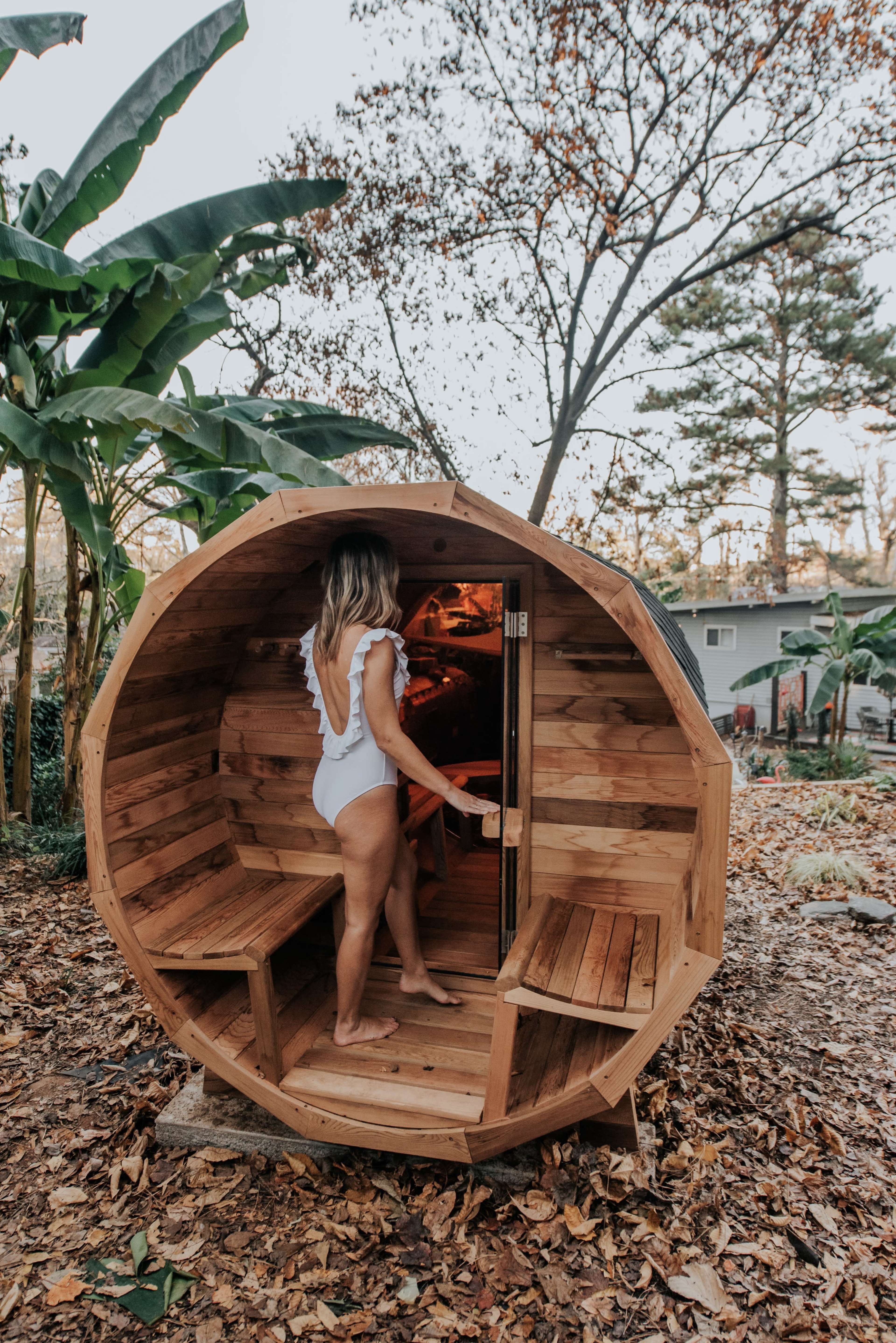 A woman in a swimsuit enters a wooden sauna nestled among tropical plants and fallen leaves.
