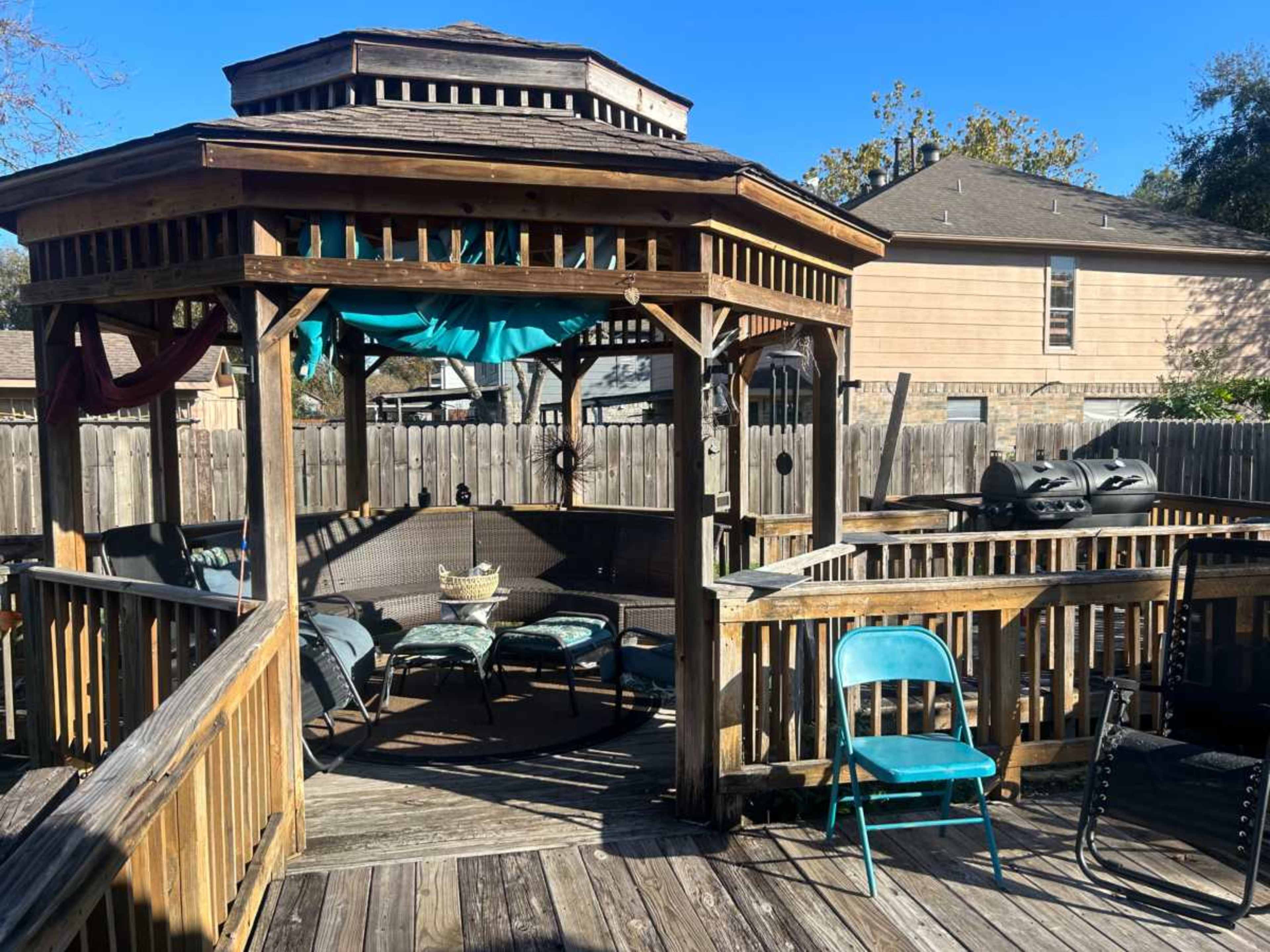 The image shows a wooden gazebo with seating inside and a few chairs around it on a deck, surrounded by a wooden fence.