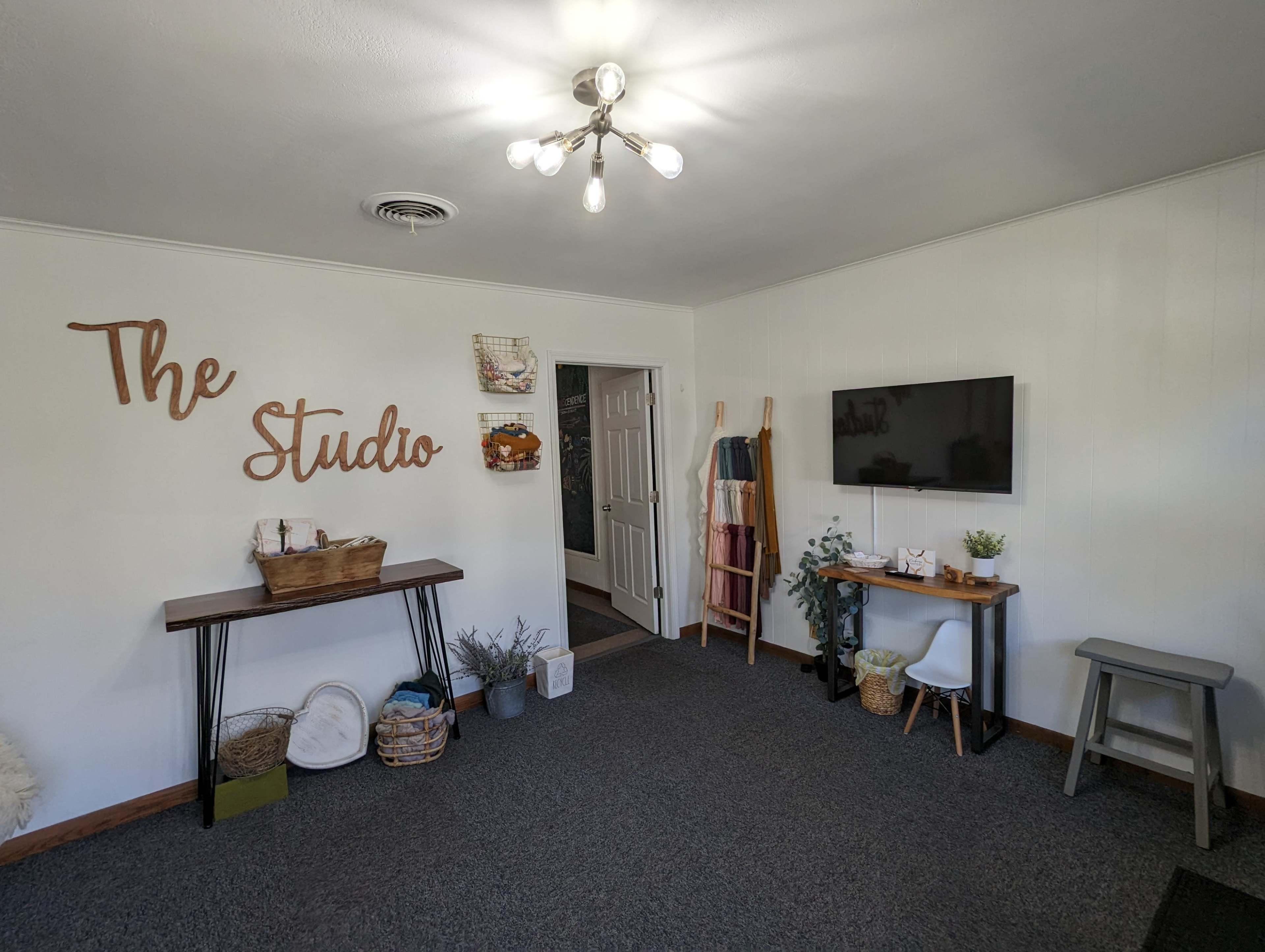 A room labeled "The Studio," featuring a wooden table, a wall-mounted television, and decorative items along with a gray stool.