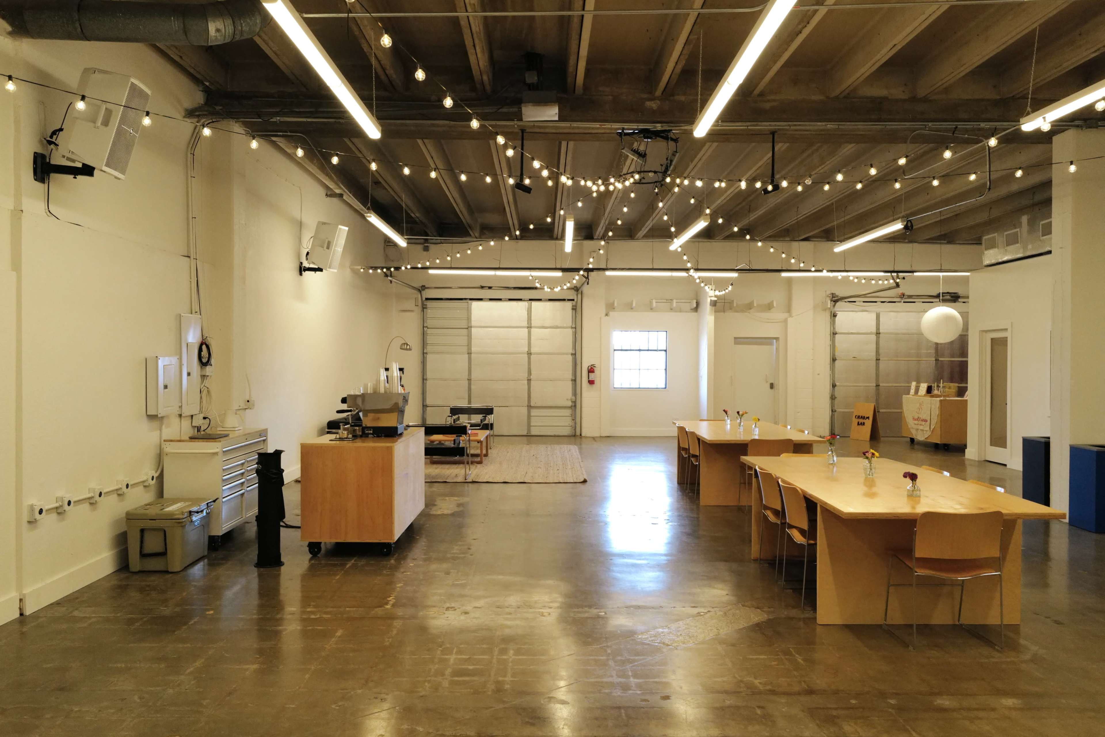 The image shows an open, spacious room with a long wooden table, a small kitchen area, and ceiling-mounted lights strung across the space.