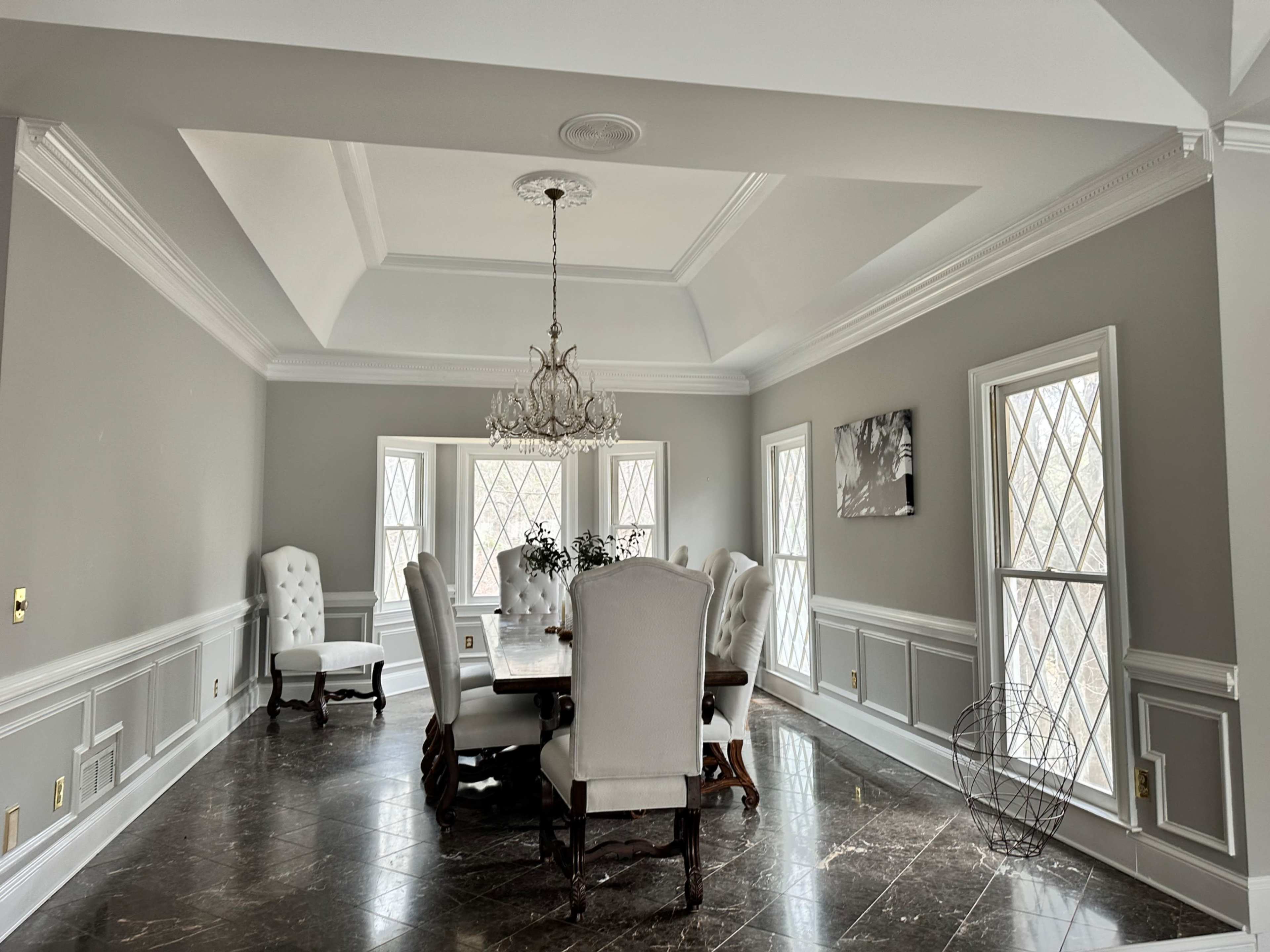 A spacious dining room features a large wooden table surrounded by six high-backed chairs, with four windows allowing natural light to enter.