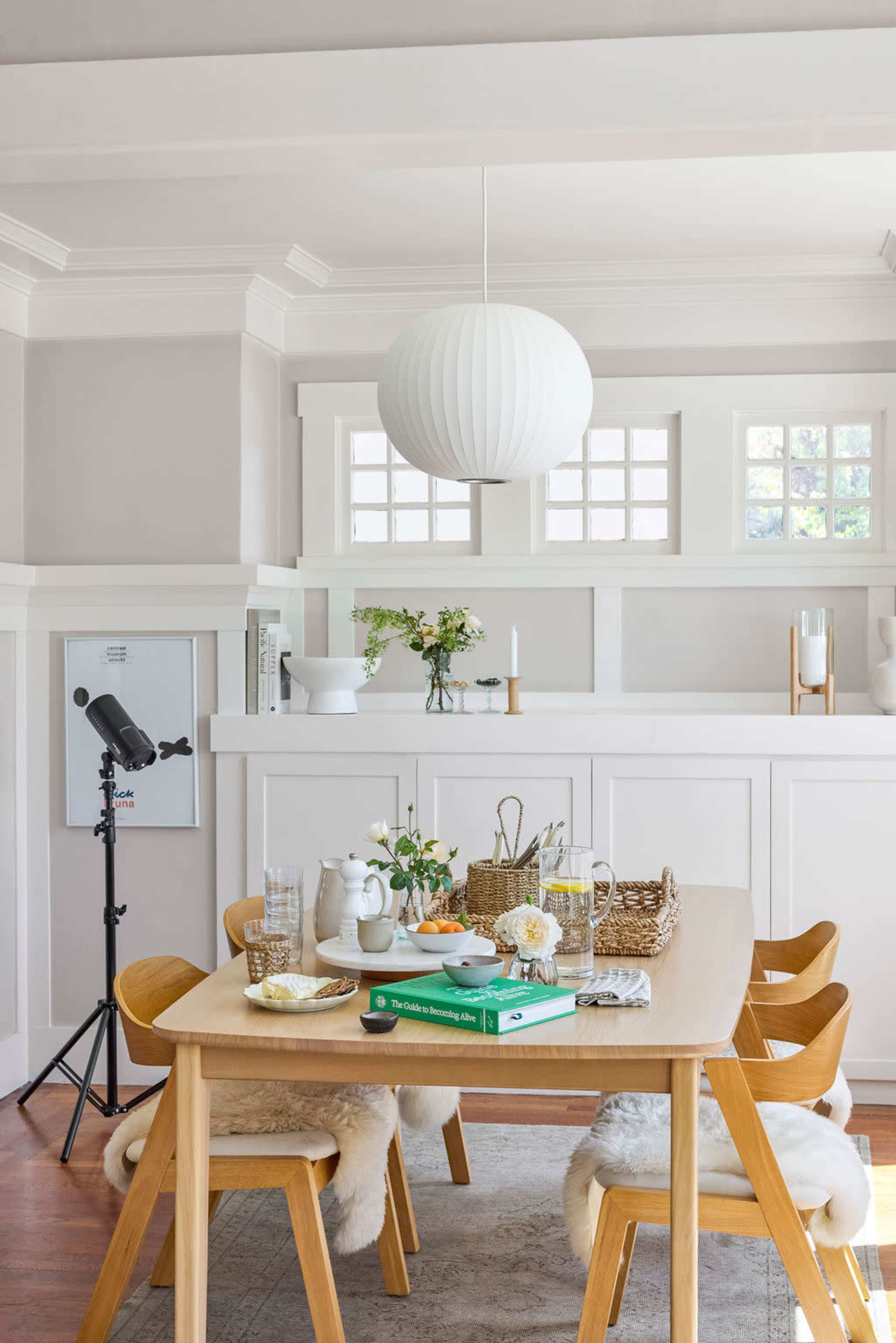 A wooden dining table set for a meal is surrounded by chairs with soft coverings, with decorative plants and objects arranged on the table and nearby shelves.
