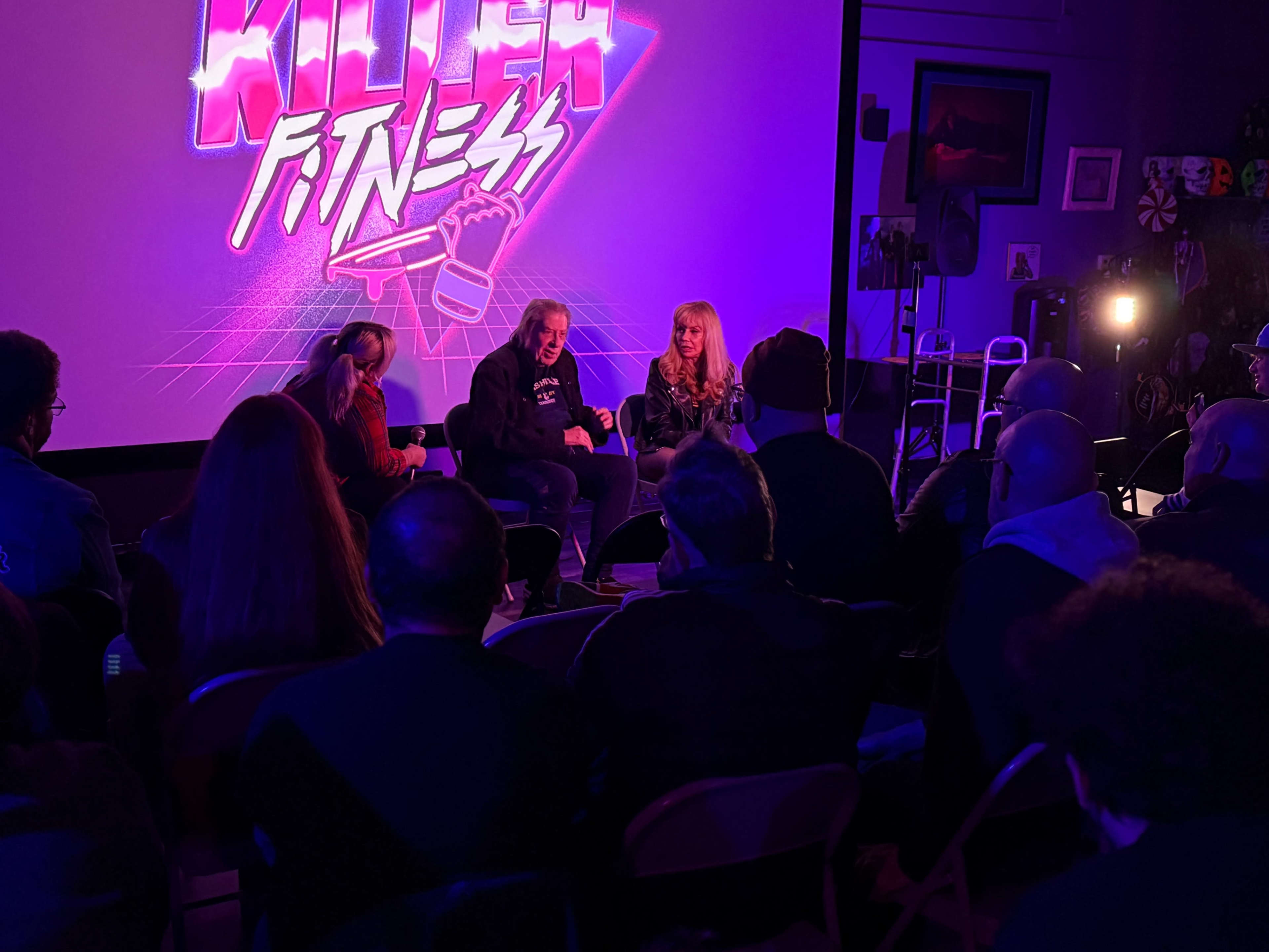 A panel discussion is taking place in a dimly lit venue, featuring three speakers in front of a large screen displaying "Killer Fitness."