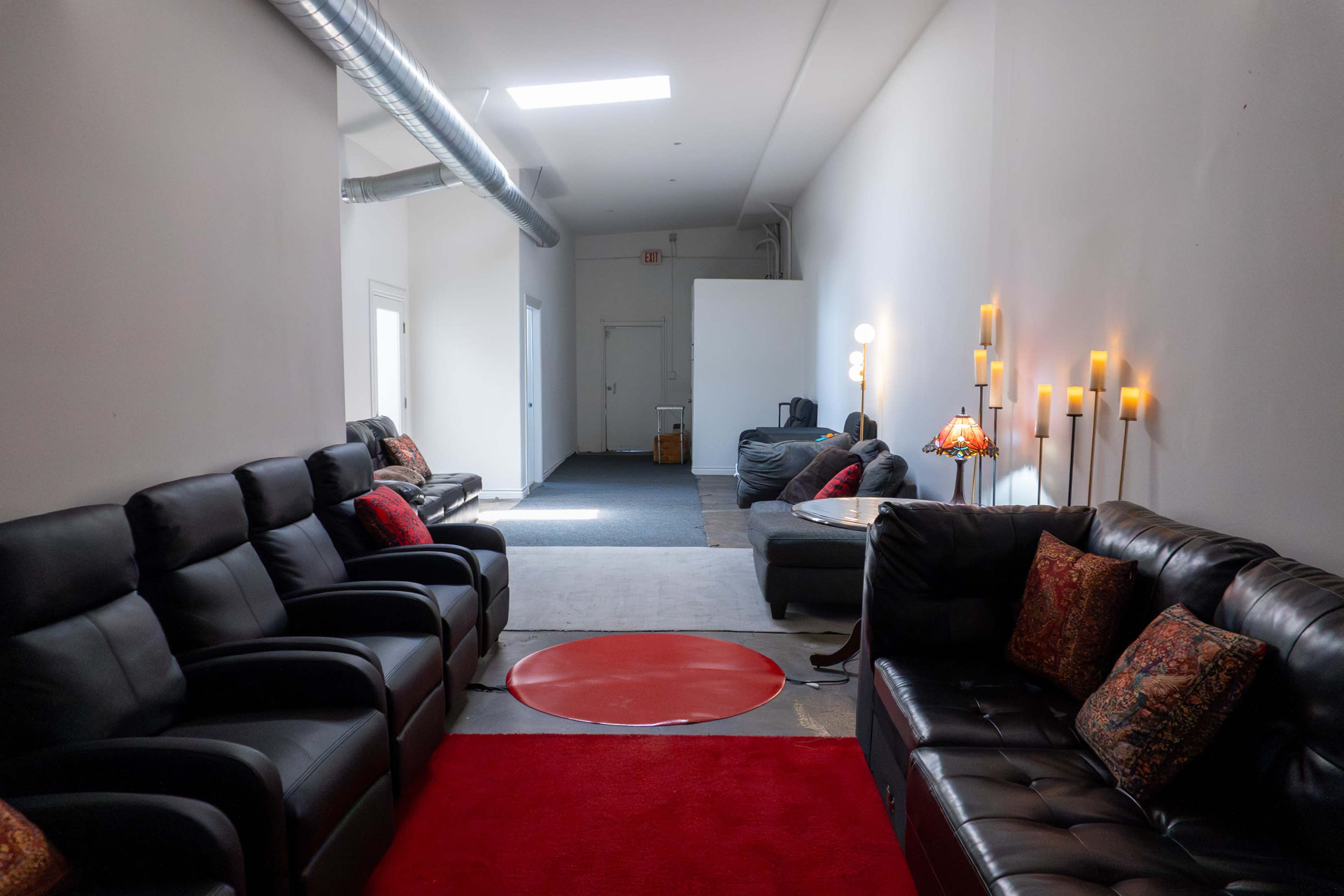 The image shows a long hallway featuring black leather sofas on either side, a red circular rug in the center, and soft lighting from wall-mounted candles.