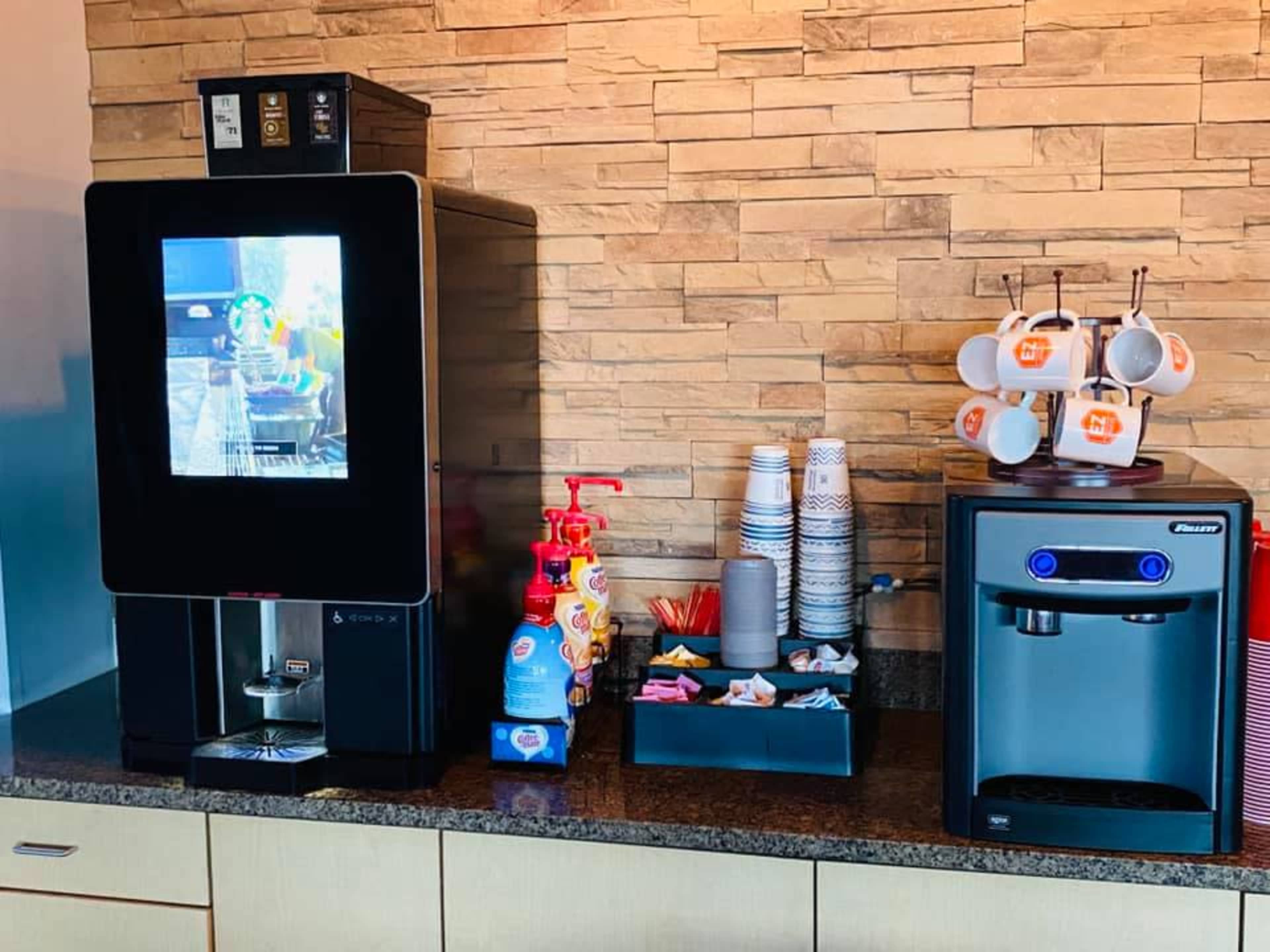 A beverage station with a coffee machine, a water dispenser, cups, condiments, and various supplies on a countertop against a stone wall.