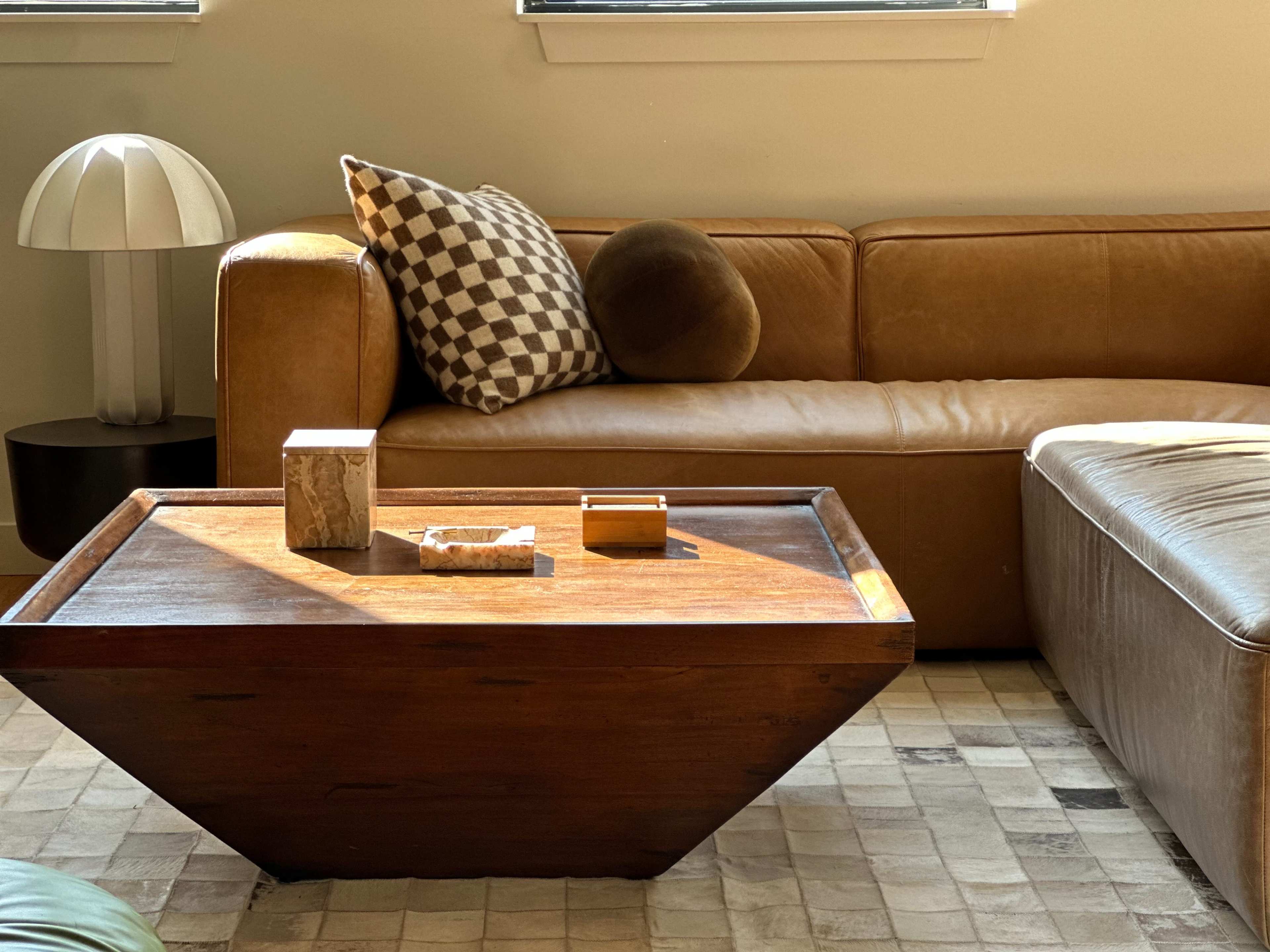 The image shows a modern living room with a brown leather sofa, a wooden coffee table, and decorative items on the table, illuminated by natural light from a nearby window.