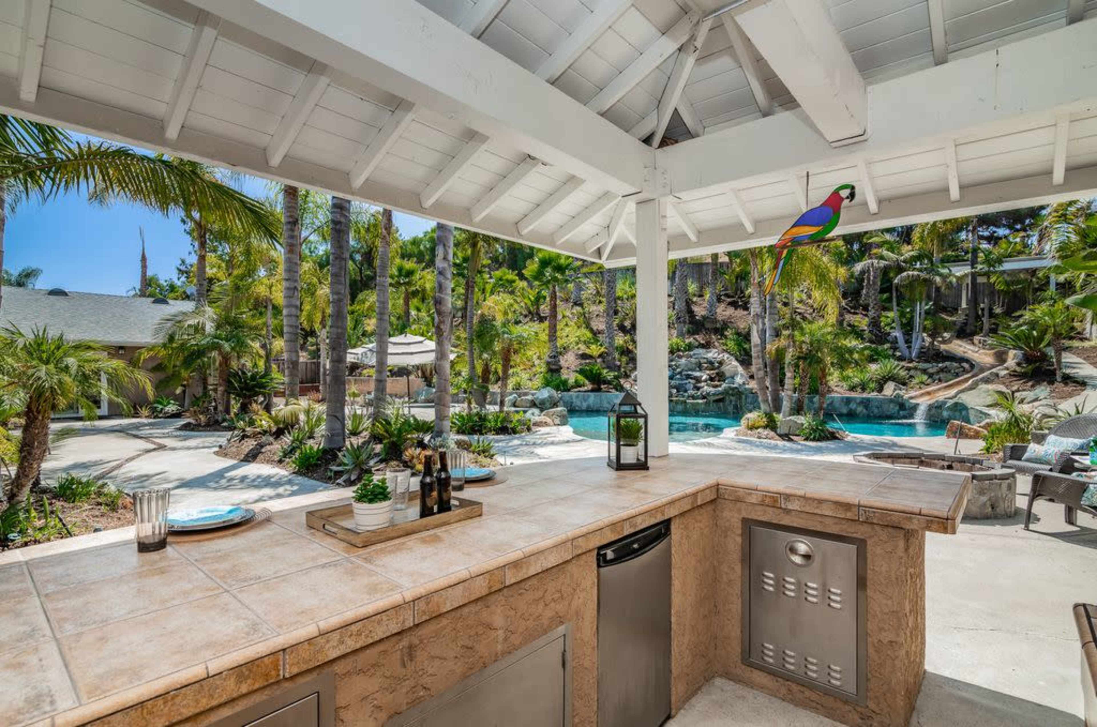 The image shows a spacious outdoor kitchen area with a countertop, sink, and grill, overlooking a landscaped backyard with a swimming pool and palm trees.