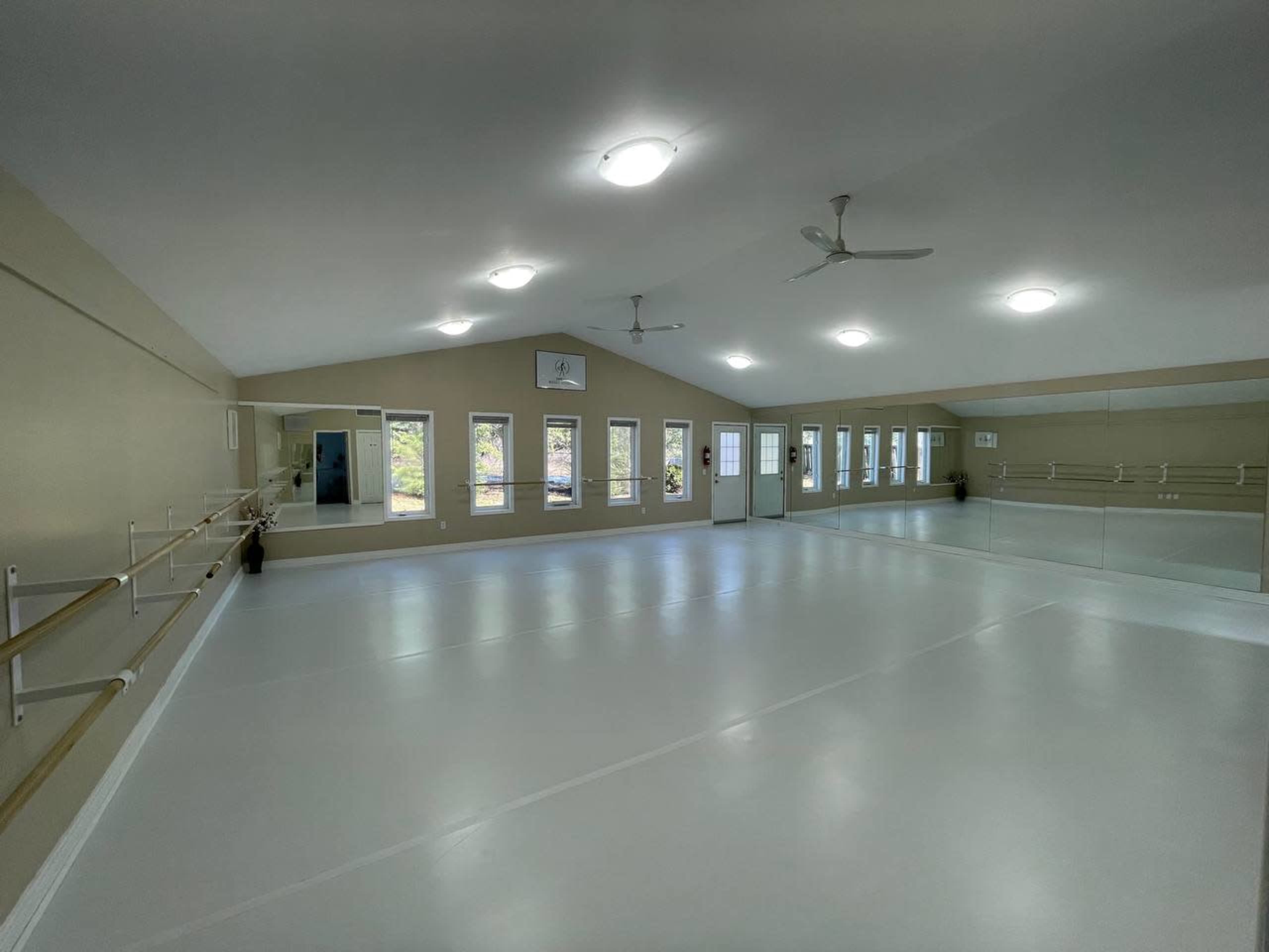 A bright dance studio features a smooth, white floor and large mirrors along one wall, with windows allowing natural light to enter.