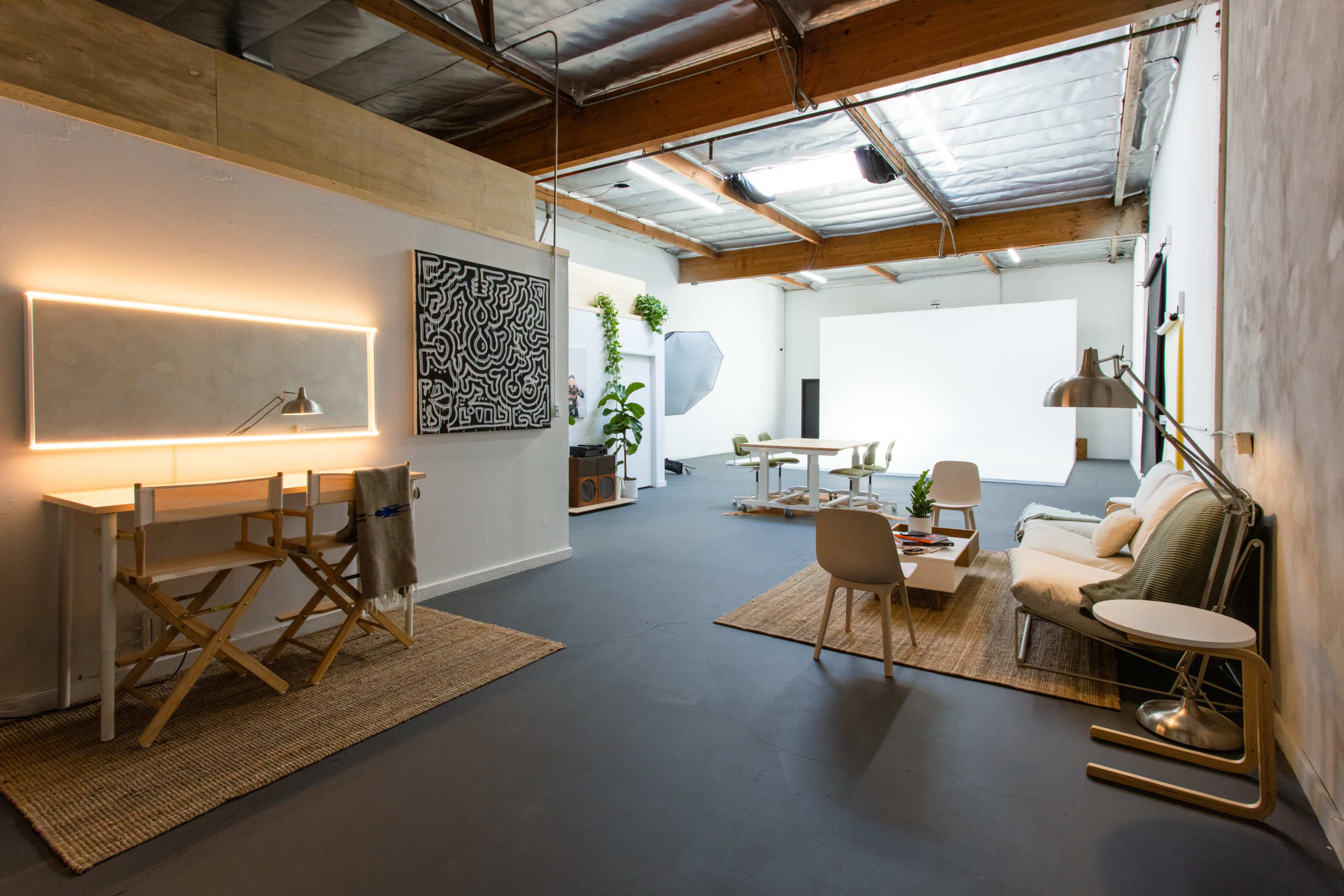 The image shows a spacious, well-lit studio featuring a seating area, a workspace with a desk and lamp, and a large blank wall suitable for presentations or photography.