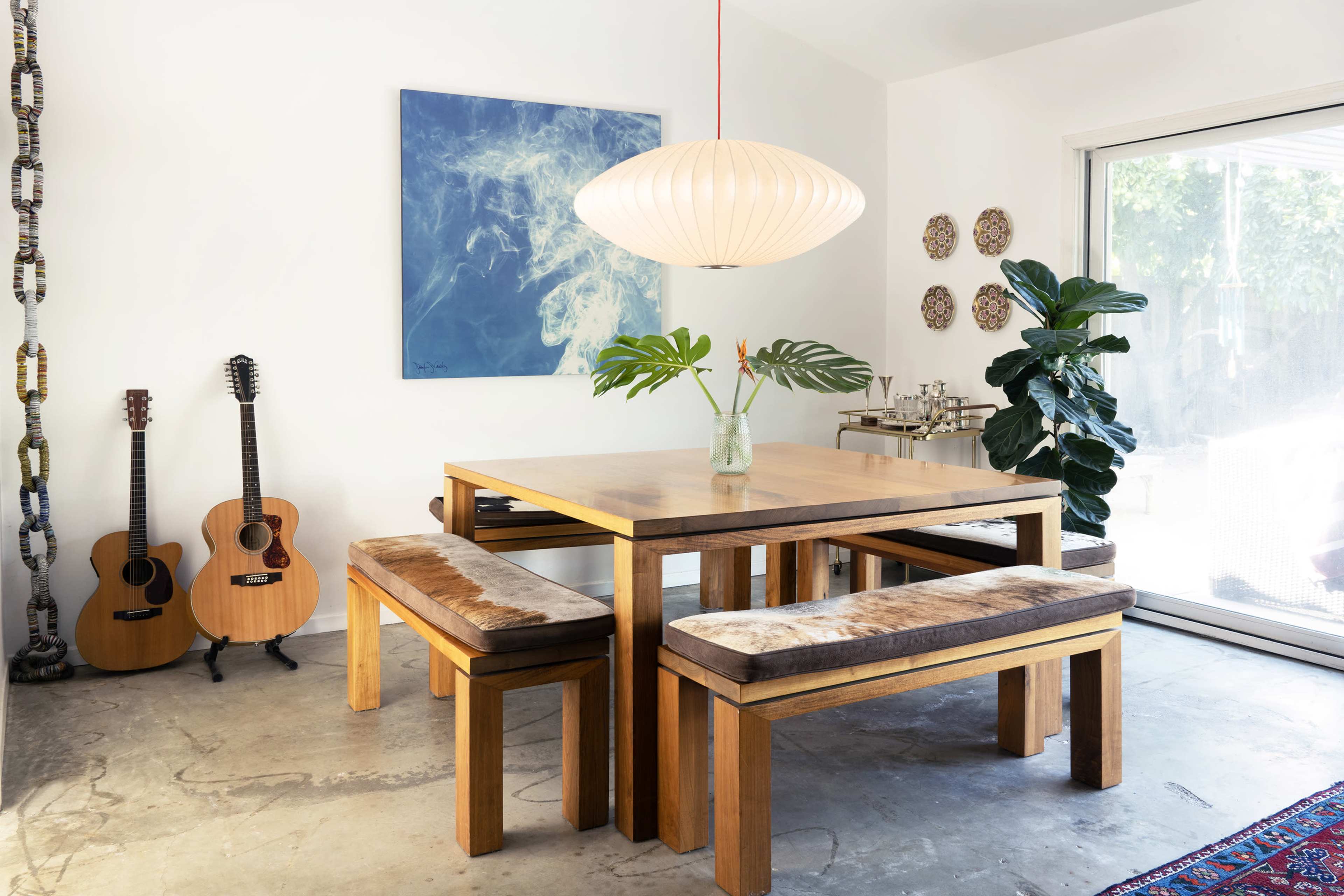 A wooden dining table with benches is positioned in a brightly lit room beside a large window, with two acoustic guitars resting against the wall and a decorative plant in a vase.
