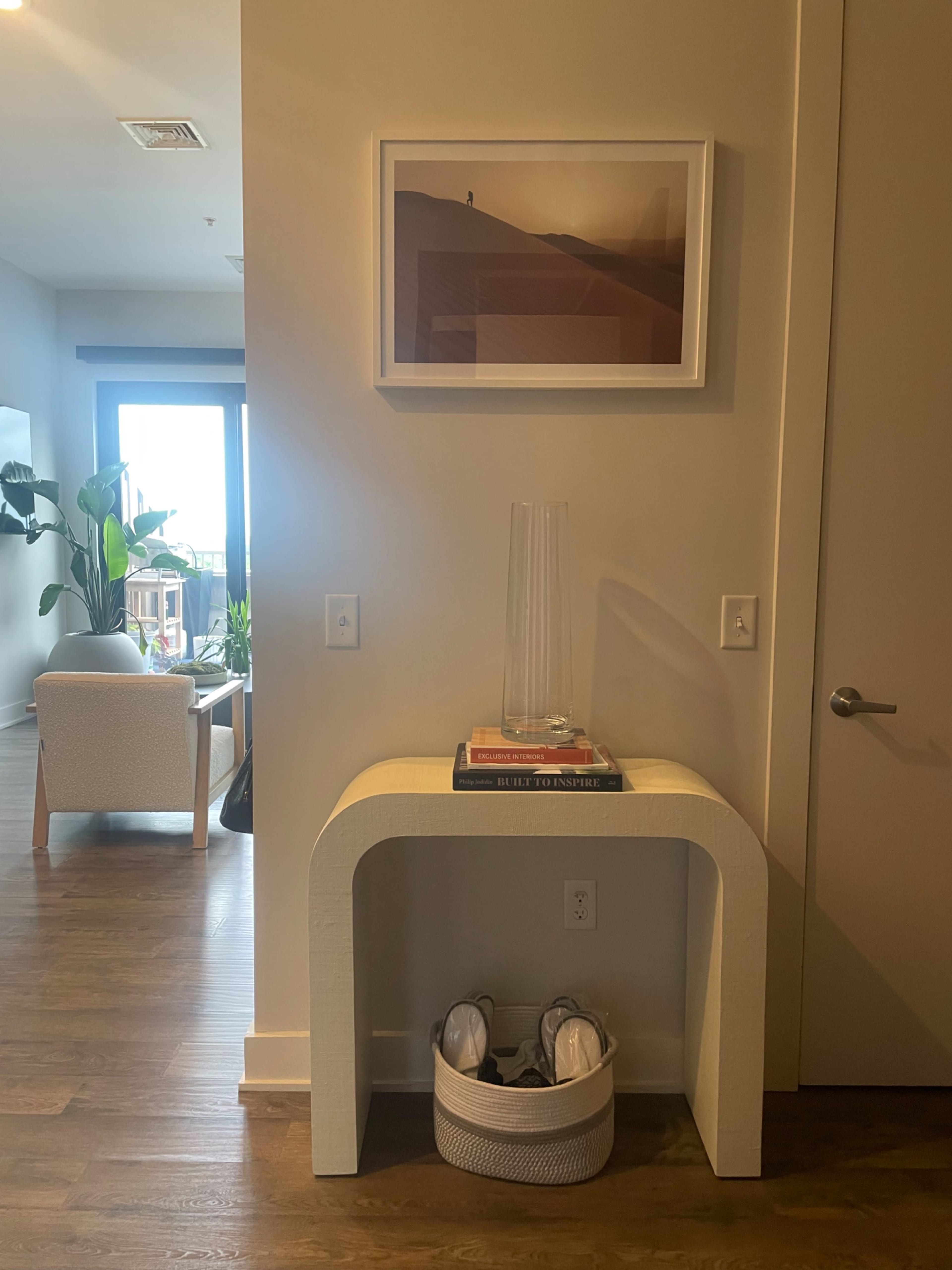 A curved white console table with a vase sits against a beige wall, with a framed artwork above it and a basket containing shoes underneath.