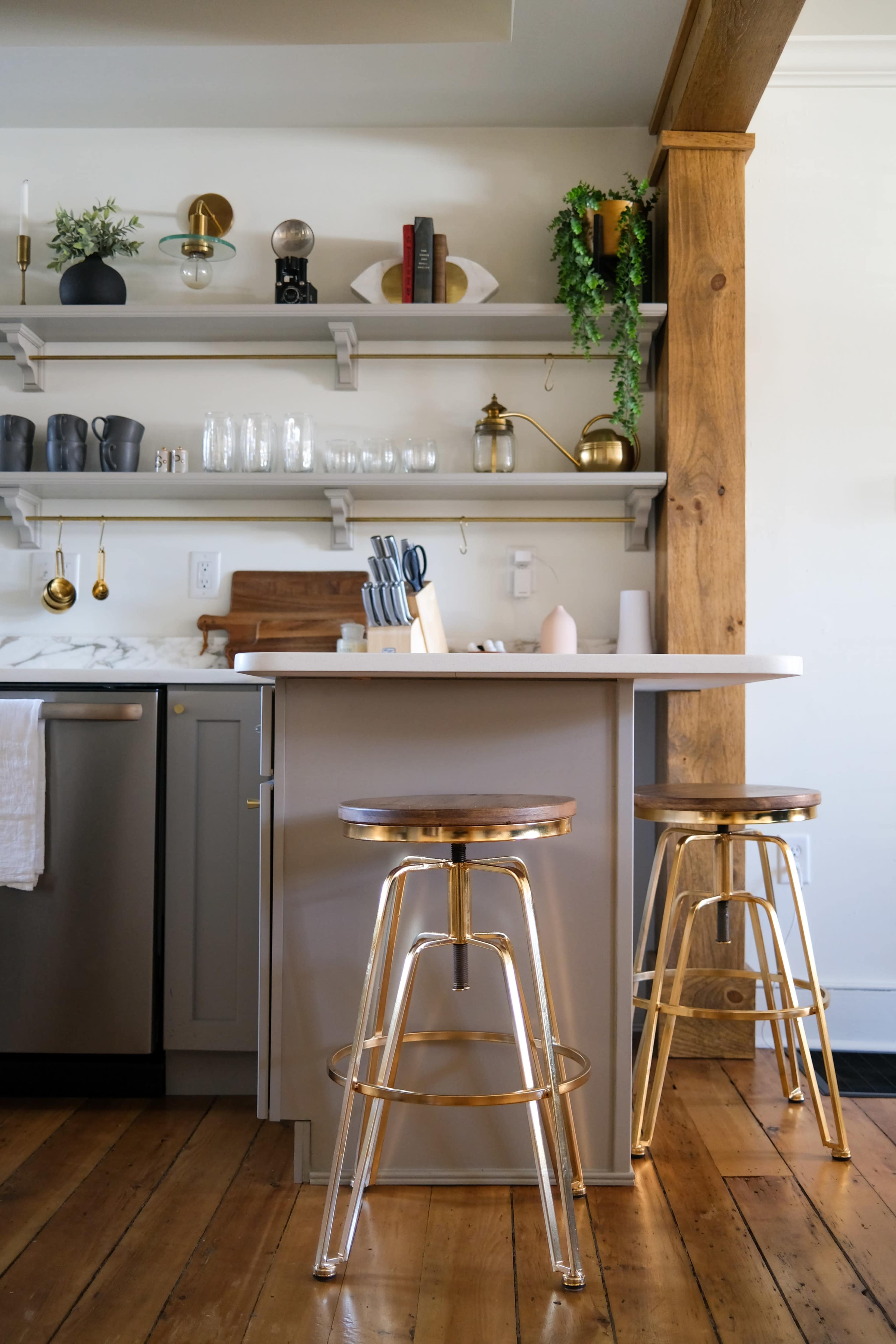 A modern kitchen features a central island with two metal stools, shelves holding kitchenware, and wooden beams on the walls.