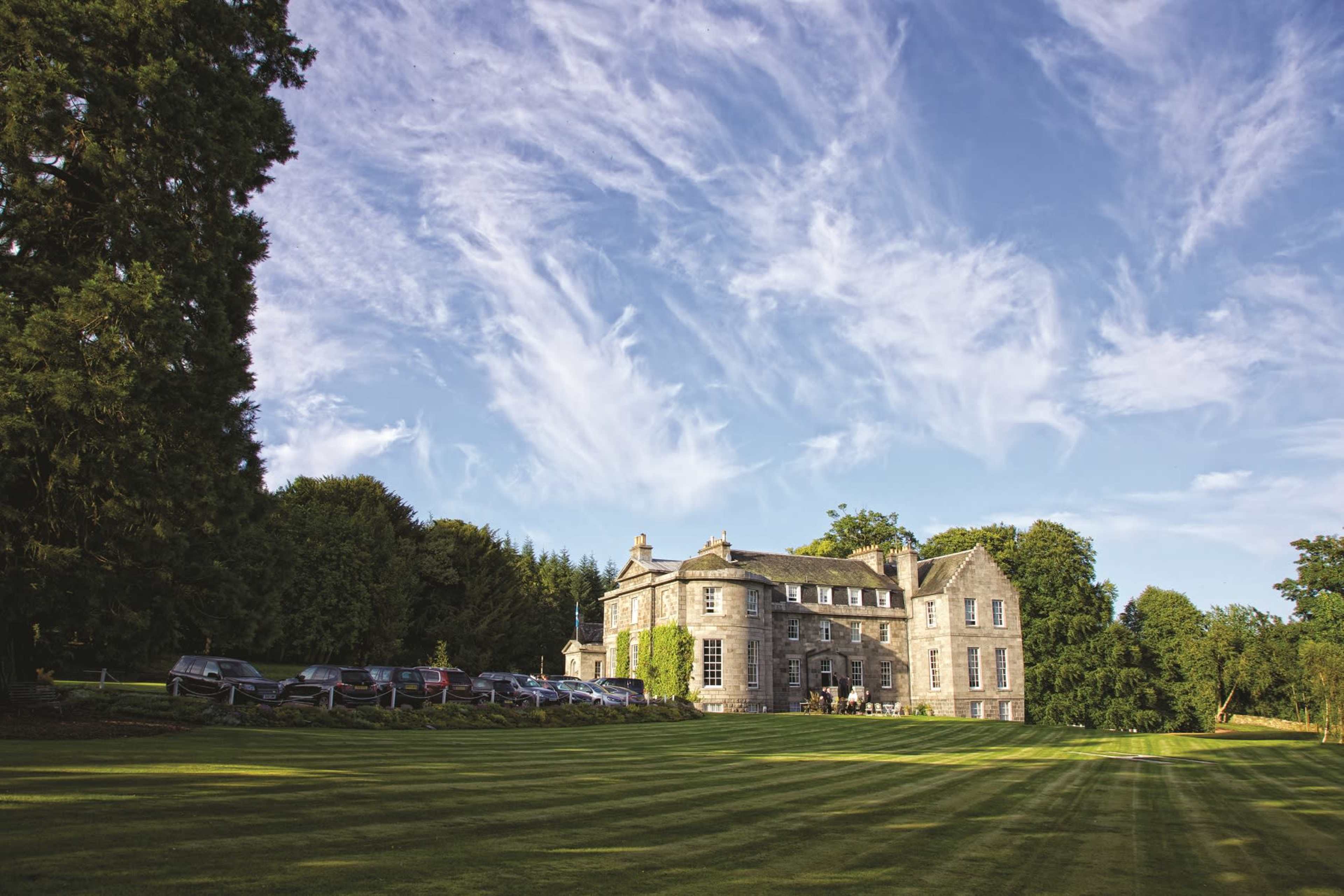 A large stone mansion with a well-maintained lawn and multiple cars parked in front under a blue sky with scattered clouds.