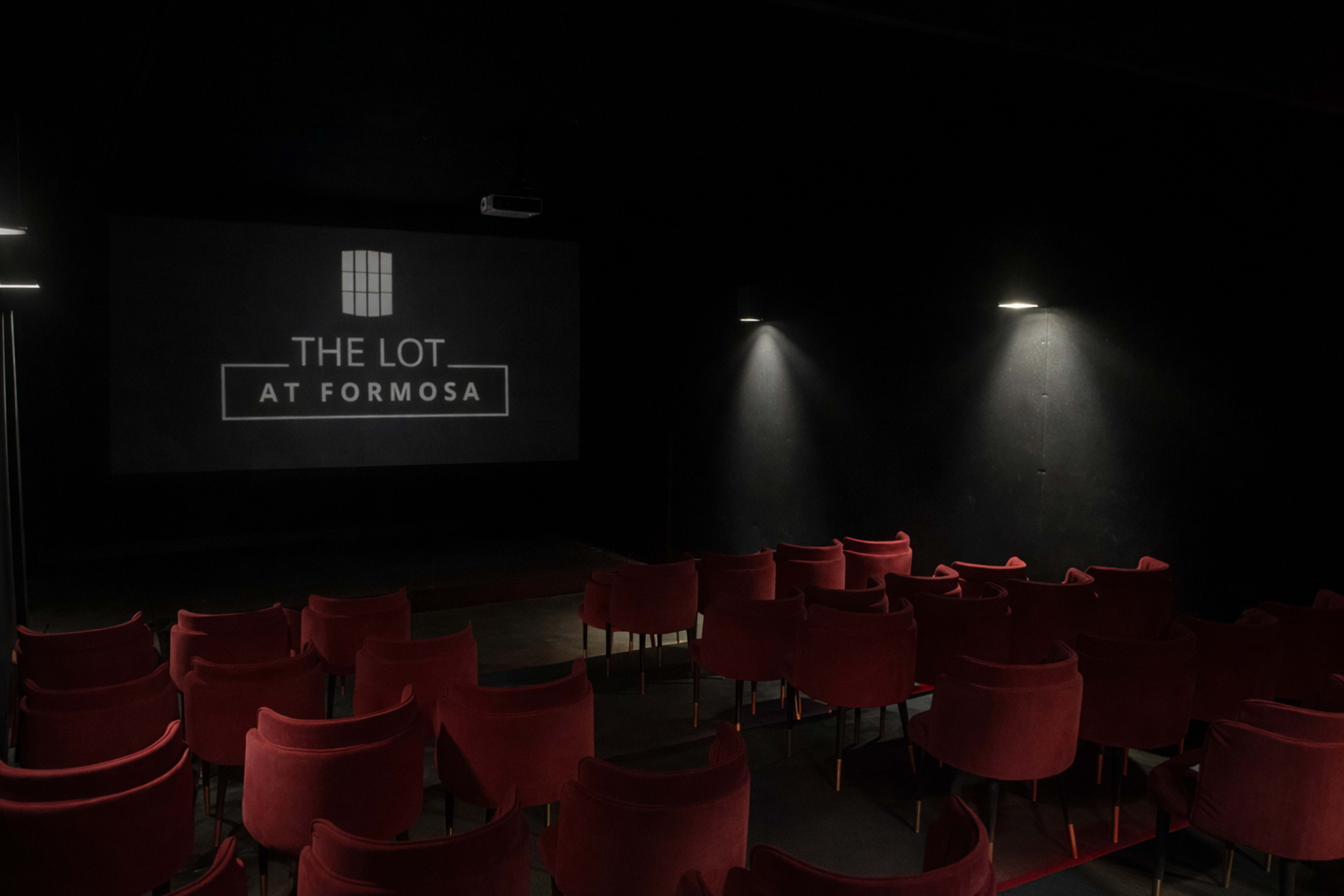 The image shows a dimly lit theater featuring red upholstered chairs arranged in a semi-circle facing a large screen displaying the text "THE LOT AT FORMOSA."