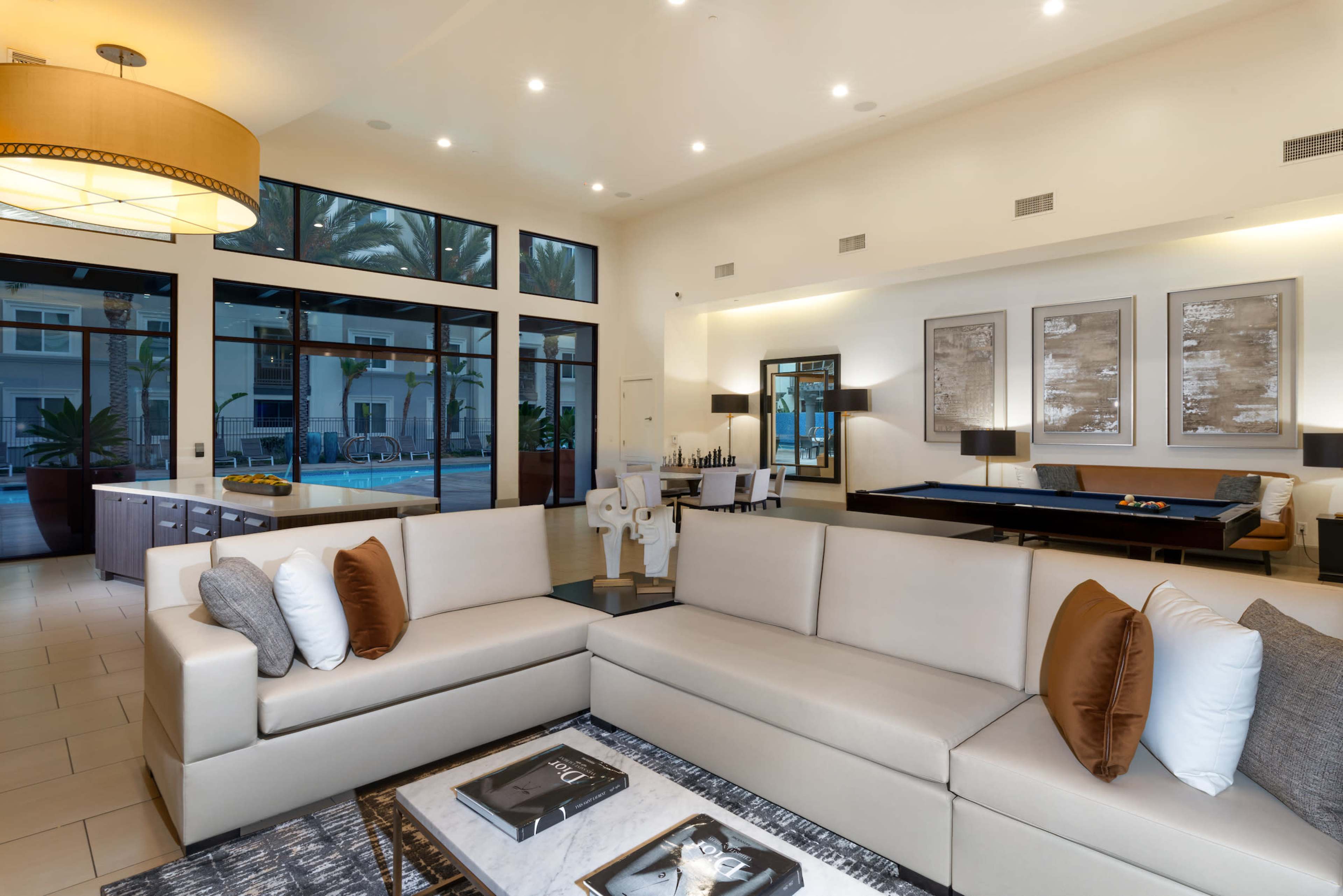 A modern living area with a light-colored sectional sofa, a coffee table with magazines, and large windows overlooking a pool area surrounded by palm trees.