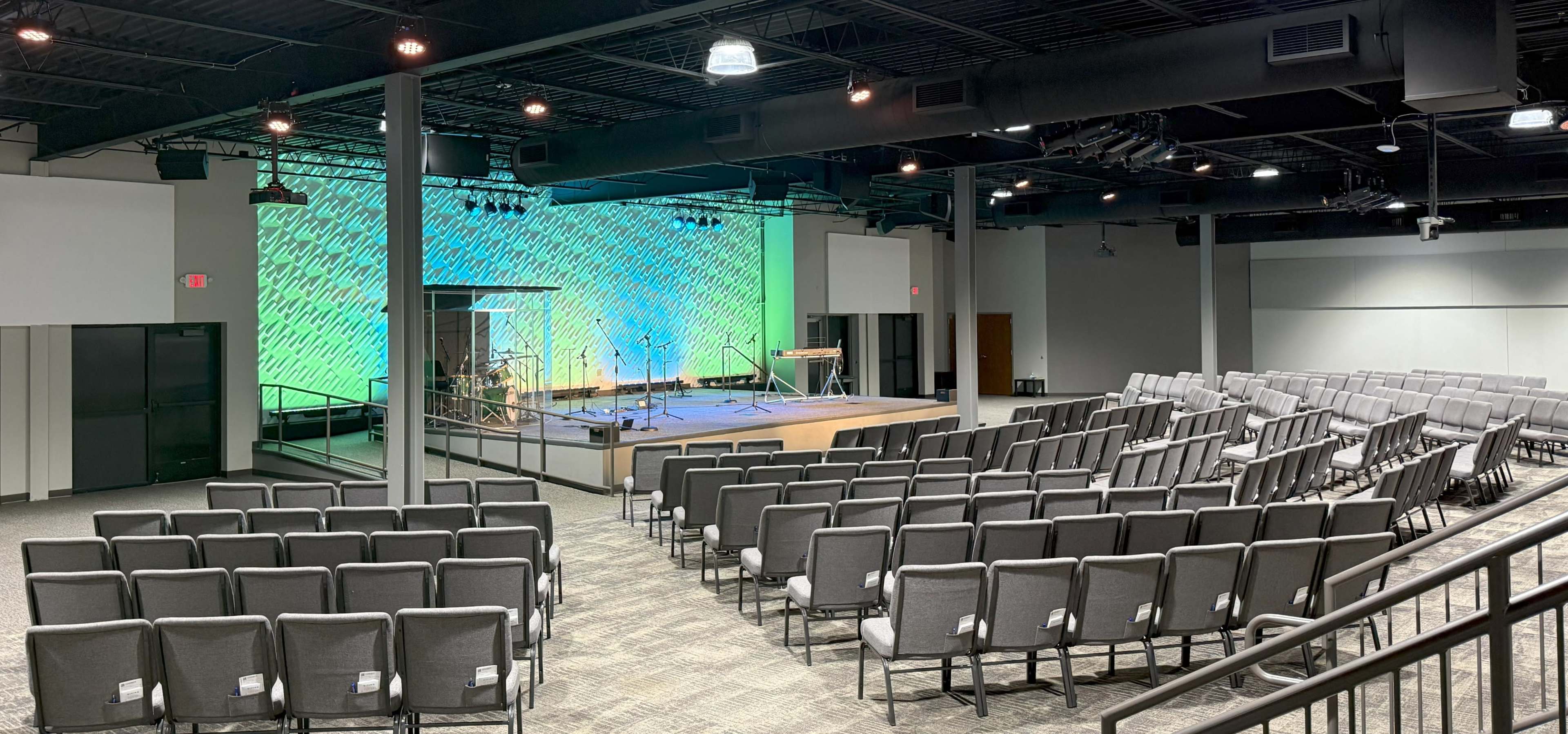 The image shows a modern auditorium setup with rows of gray chairs facing a stage equipped with musical instruments and a colorful backdrop.