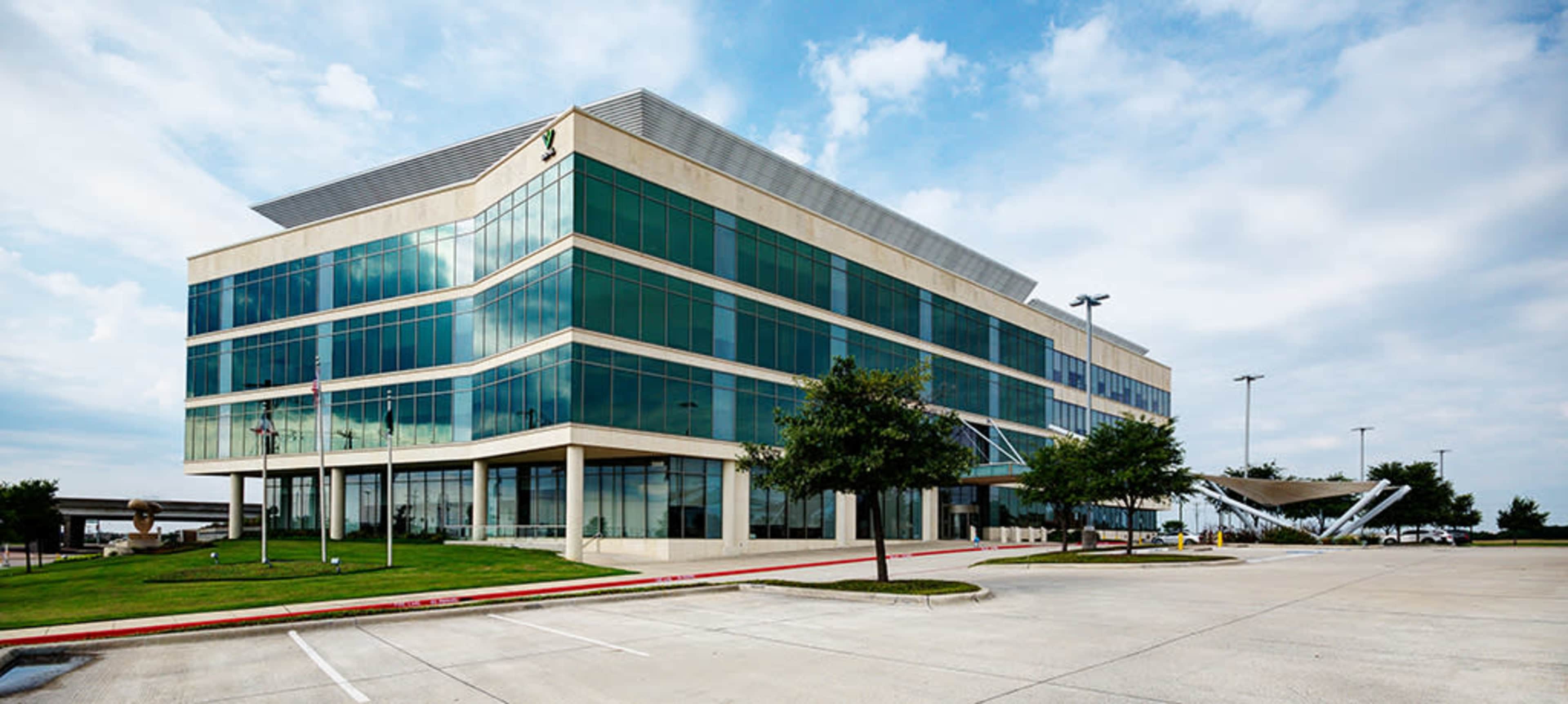 A modern, multi-story office building with large glass windows is situated in a parking lot under a cloudy sky.