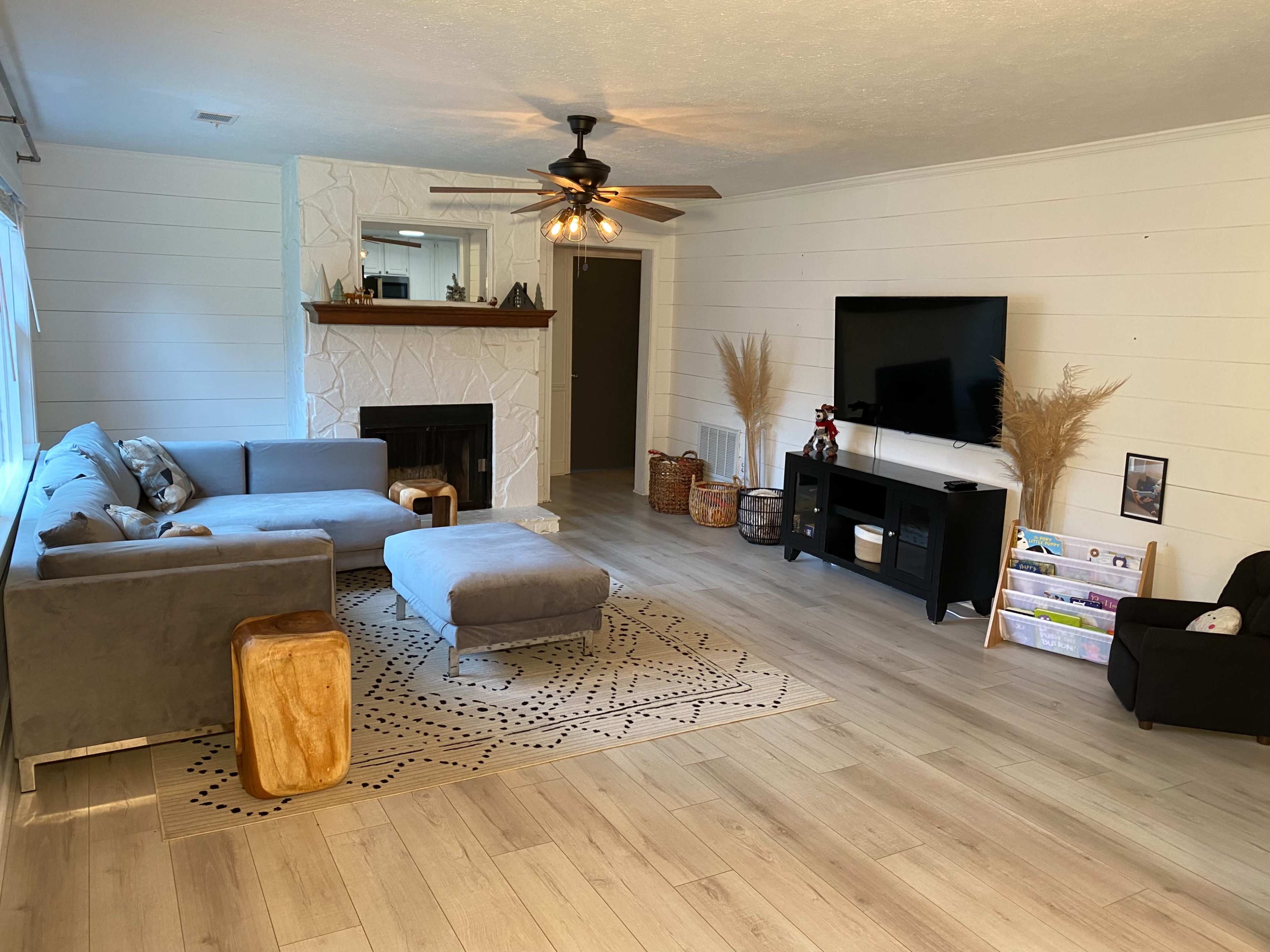 The living room features a gray sectional sofa, a fireplace with a wooden mantel, a television mounted on a wall, and light wooden flooring.