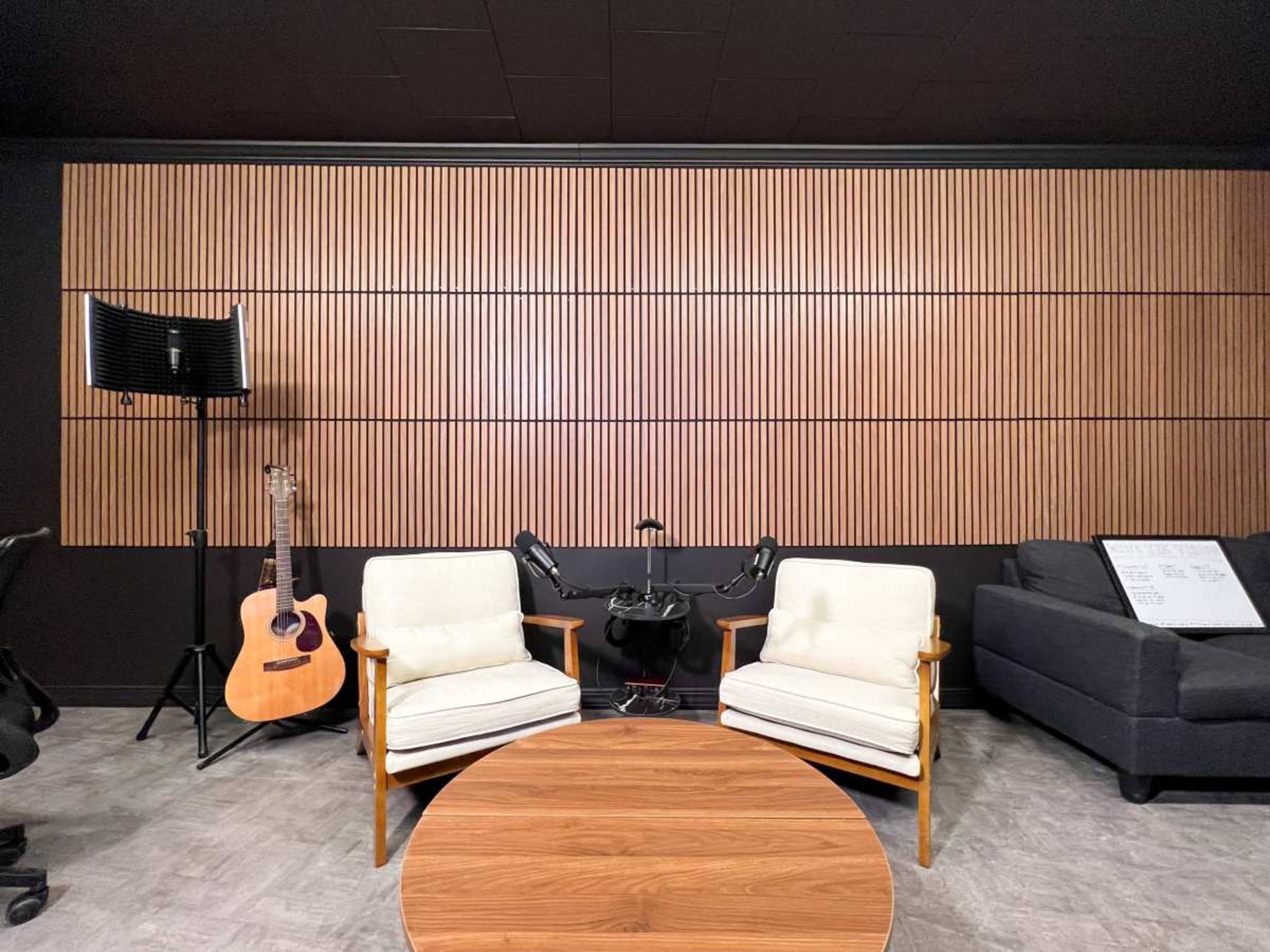The image features a modern interior space with two white armchairs, a wooden coffee table, an acoustic guitar on a stand, and a dark-colored couch against a wood-paneled wall.
