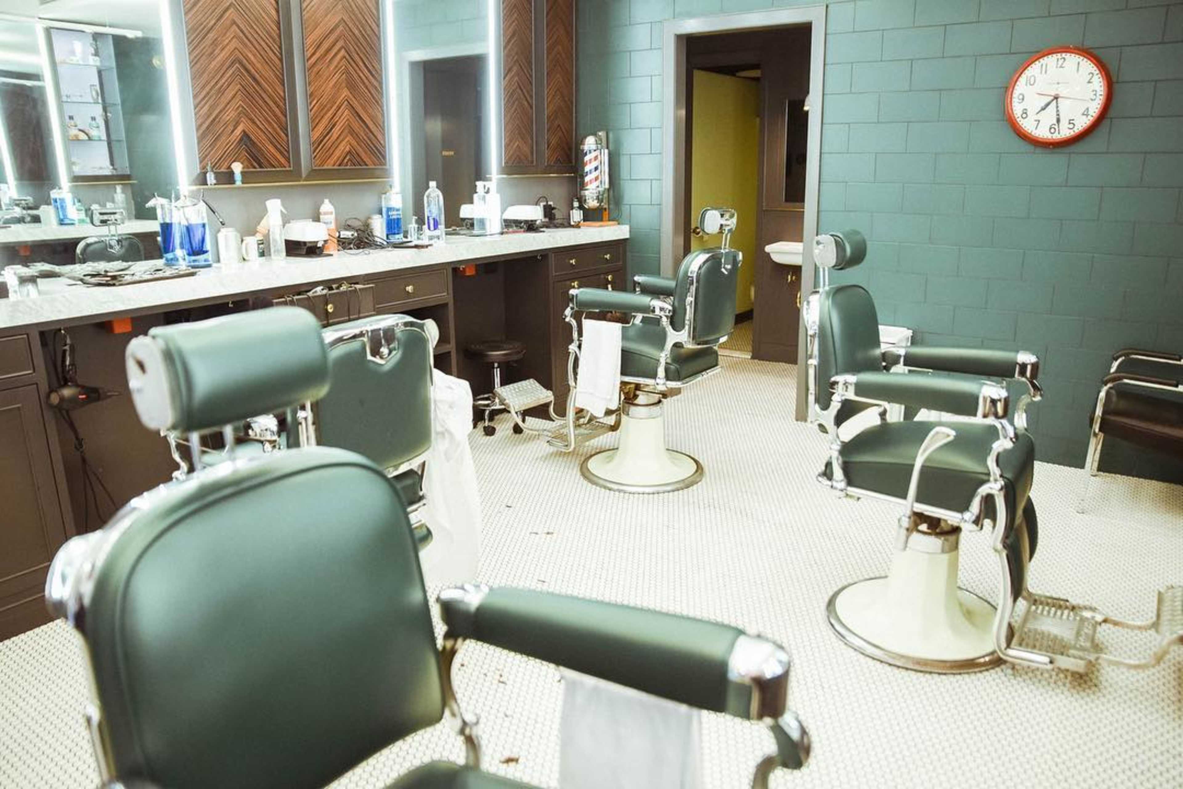 A barber shop interior with multiple green and silver barber chairs, a counter with various grooming products, and a wall clock.