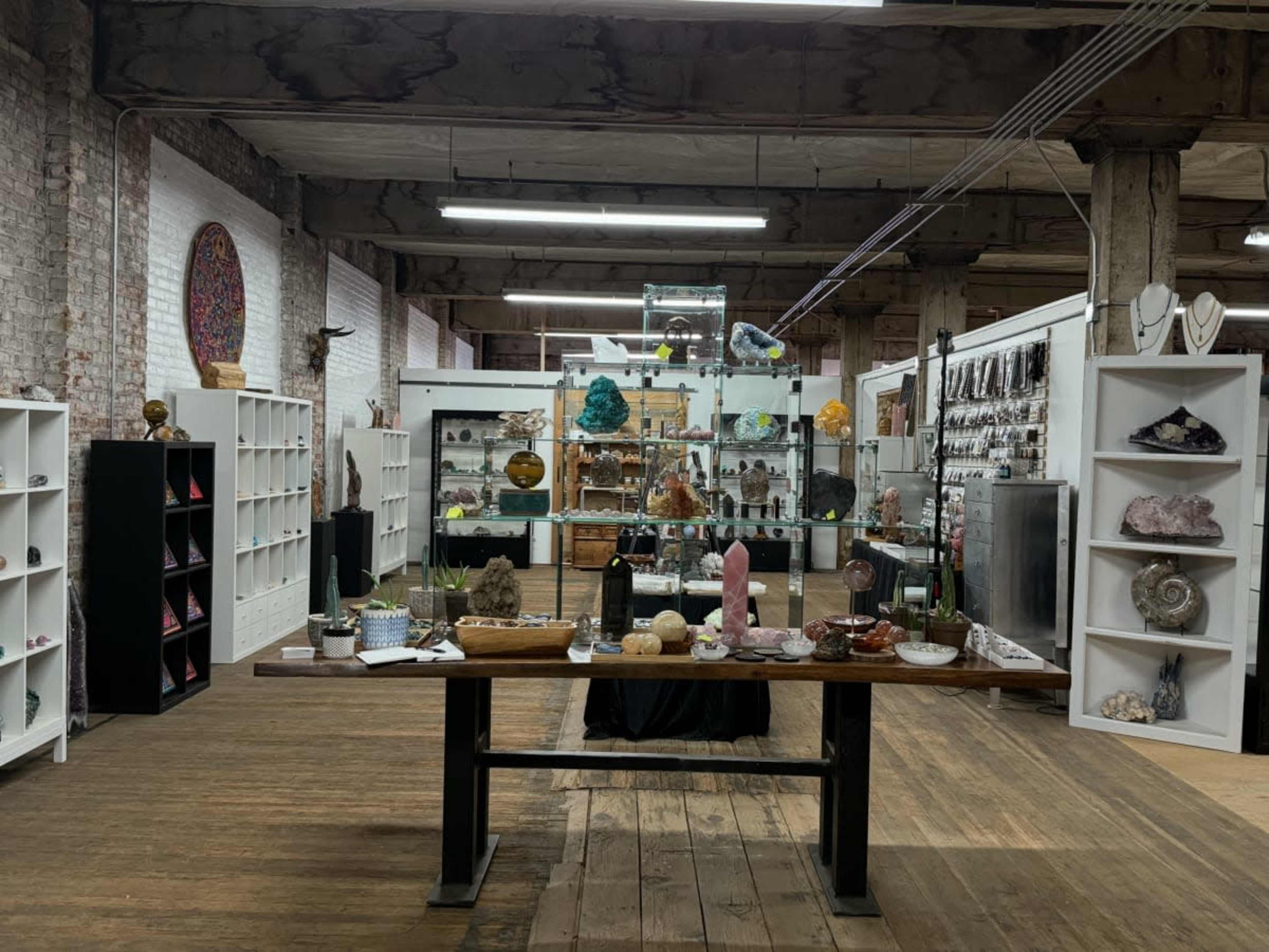The image shows a spacious store interior featuring displays of various crystals and minerals on shelves and tables, with exposed brick walls and wooden flooring.