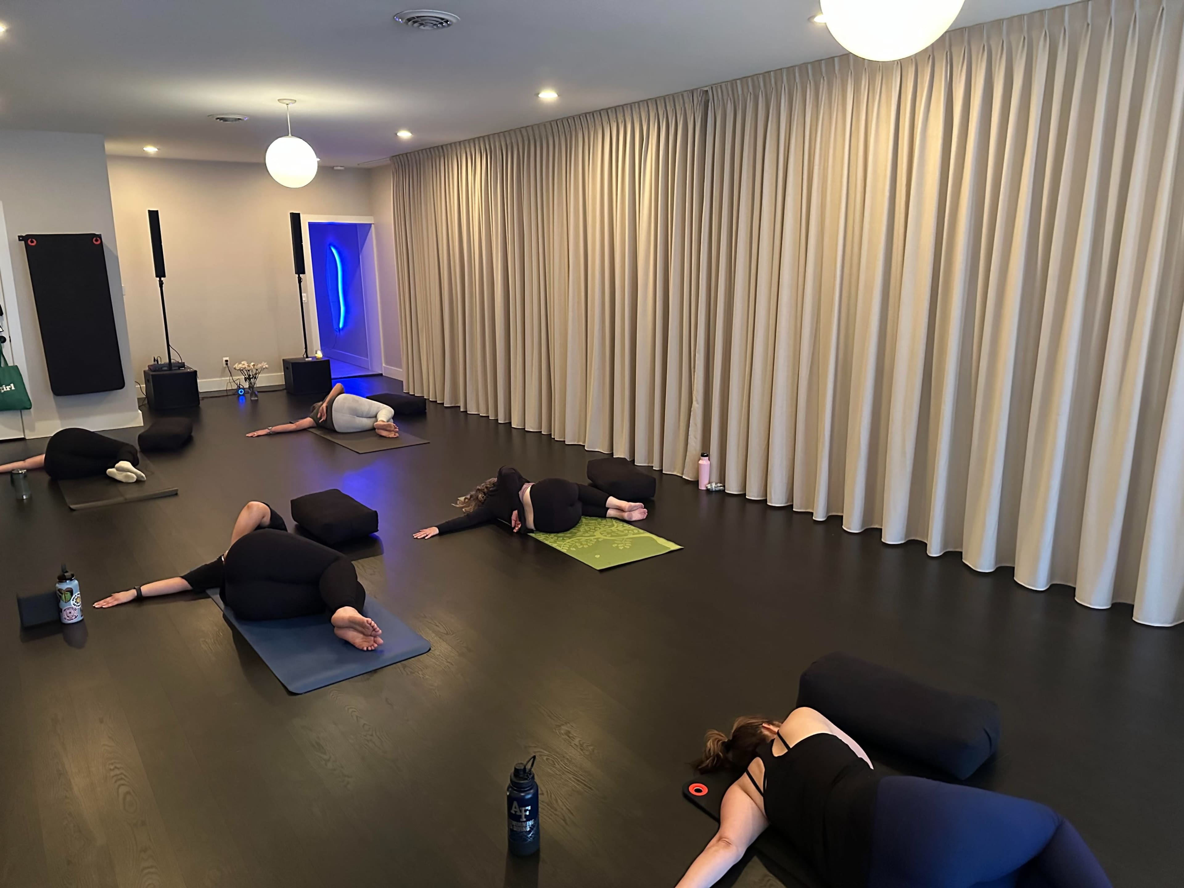 A group of individuals practice a yoga session on mats in a studio with soft lighting and beige curtains.